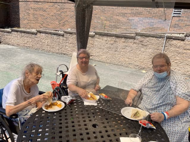 Three elderly individuals sitting around a black metal outdoor table under a shaded area, eating food from white plates. Two women and one man are present, with one woman and the man in wheelchairs. The man is wearing a hospital gown and a face mask pulled down below his chin. A brick wall and pavement are visible in the background.