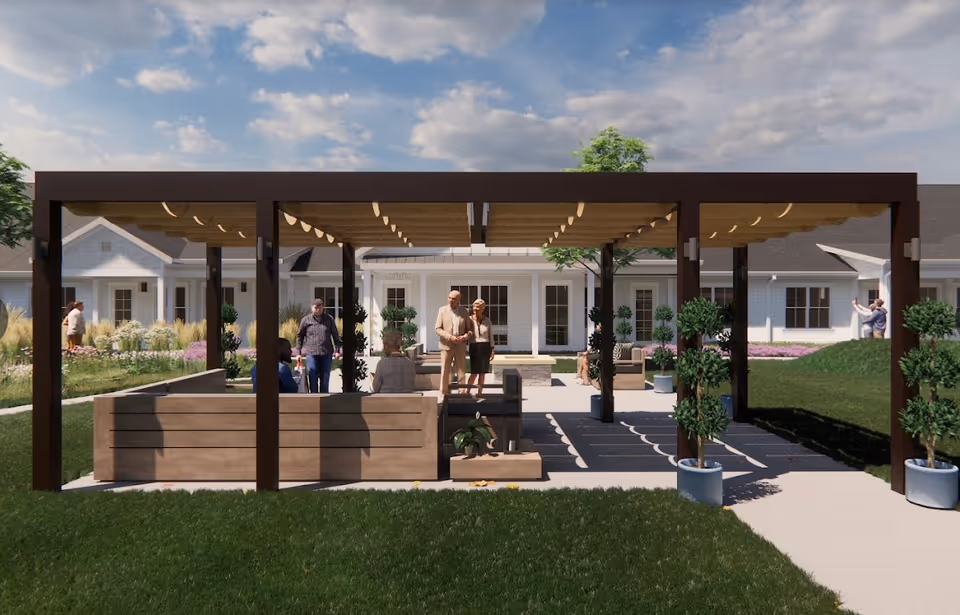 Outdoor seating area with a pergola structure providing shade, surrounded by potted plants and greenery. Several people are seen socializing and relaxing in the space, with a senior living facility building in the background under a partly cloudy sky.