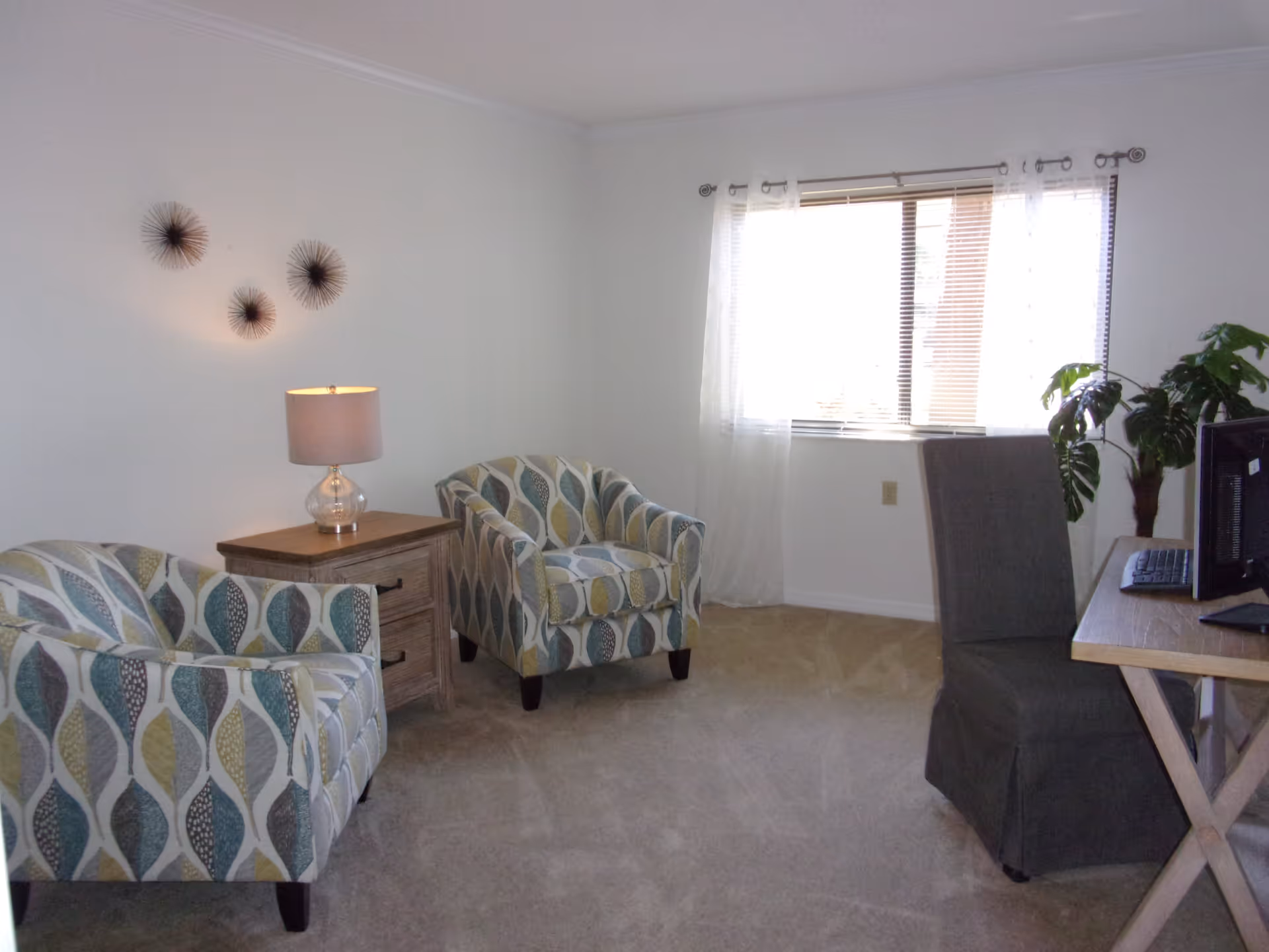 A bright room with two patterned armchairs, a wooden side table with a lamp, a window with sheer white curtains, a gray upholstered chair, a wooden desk with a computer monitor and keyboard, and a green potted plant in the corner.