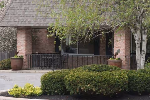 Brick building exterior with a front porch, railing, and potted plants.