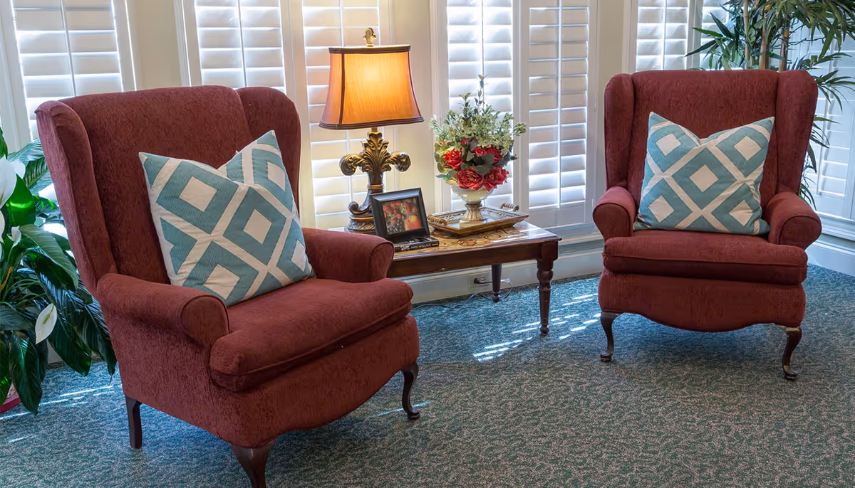Two red upholstered armchairs with blue and white patterned pillows sit on a green carpeted floor in front of white plantation shutters. Between the chairs is a wooden side table with a decorative lamp, a framed photo, and a floral arrangement. There are green plants on either side of the chairs.