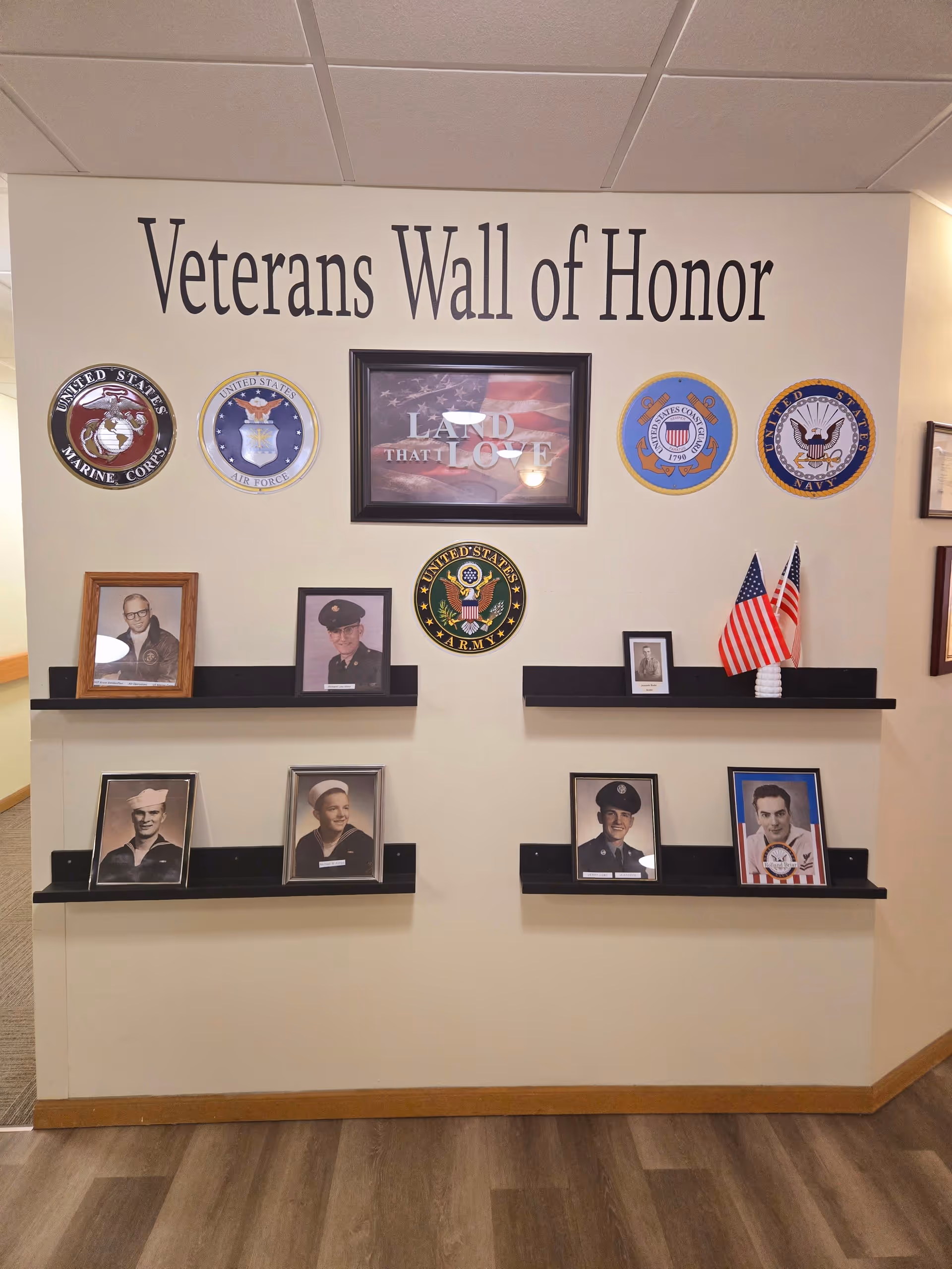 A Veterans Wall of Honor display with framed photos of veterans, military branch emblems, and two small American flags on black shelves mounted on a beige wall. The text 'Veterans Wall of Honor' is written above the display.