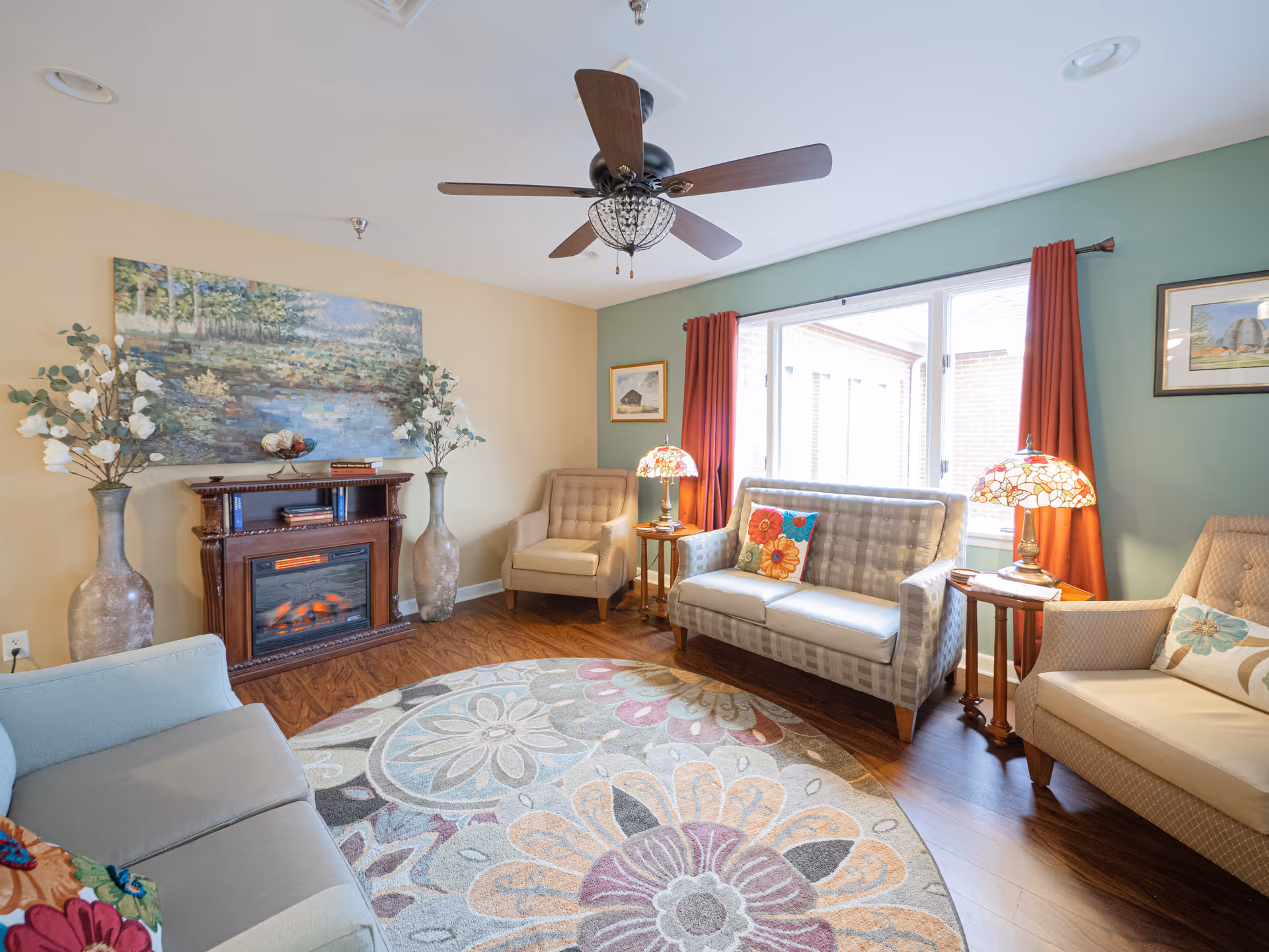 A cozy living room with a ceiling fan, a fireplace with decorative vases on either side, a large floral painting above the fireplace, a patterned round rug, two armchairs, a loveseat with colorful pillows, two side tables with stained glass lamps, and large windows with red curtains.