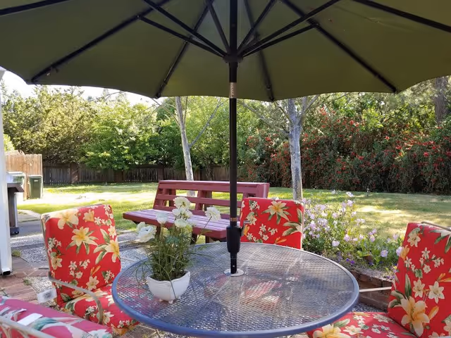 Outdoor patio area with a round metal table and four chairs with red floral cushions. A large green umbrella is centered over the table. There is a white flower pot with white flowers on the table. In the background, there is a red wooden bench, green grass, trees, and bushes with red flowers.