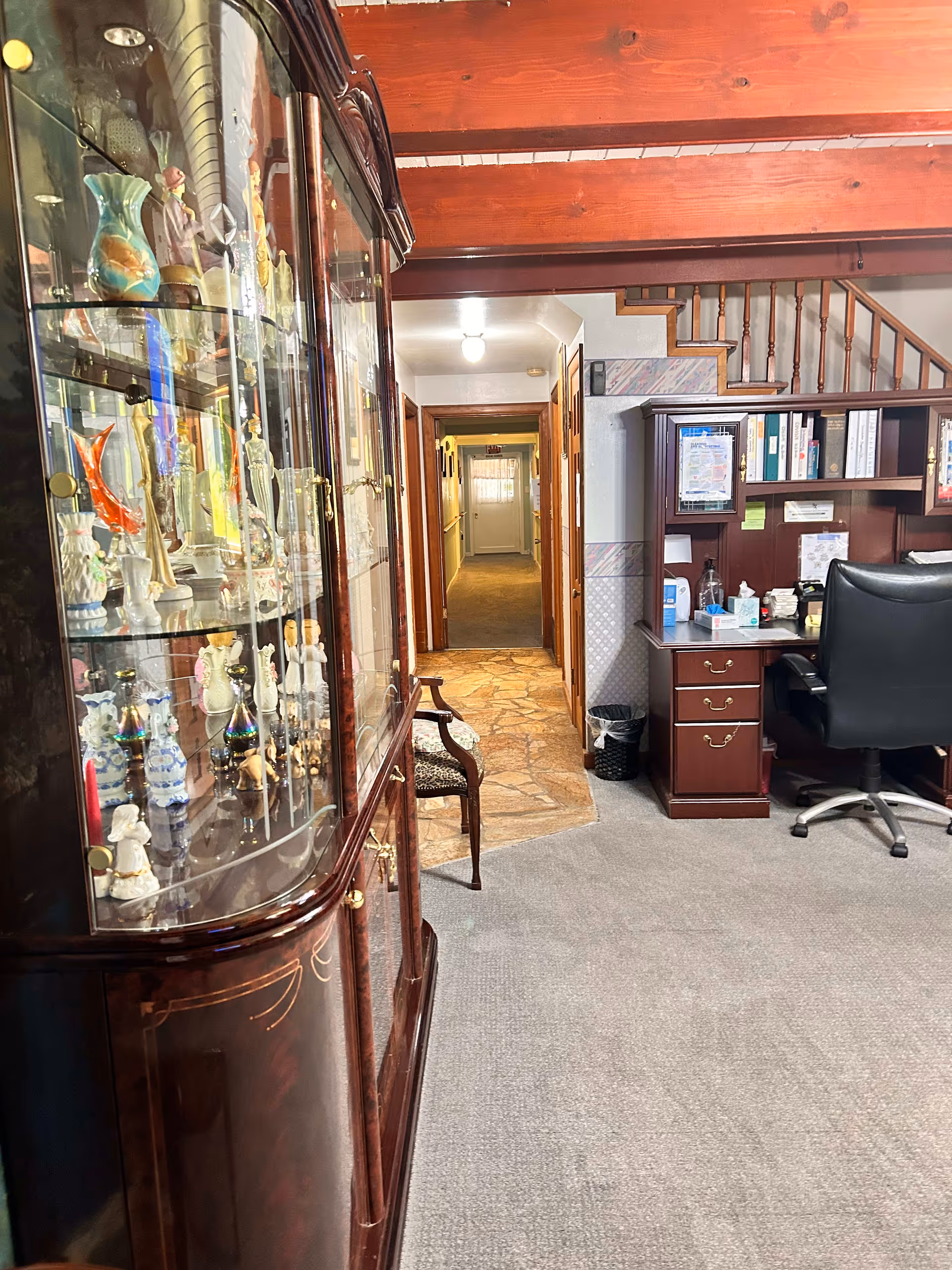 Interior view of a senior living facility showing a hallway with a wooden display cabinet filled with decorative items on the left, a wooden desk with office supplies and a black office chair on the right, and a staircase in the background. The floor is carpeted with a stone tile section near the hallway entrance.