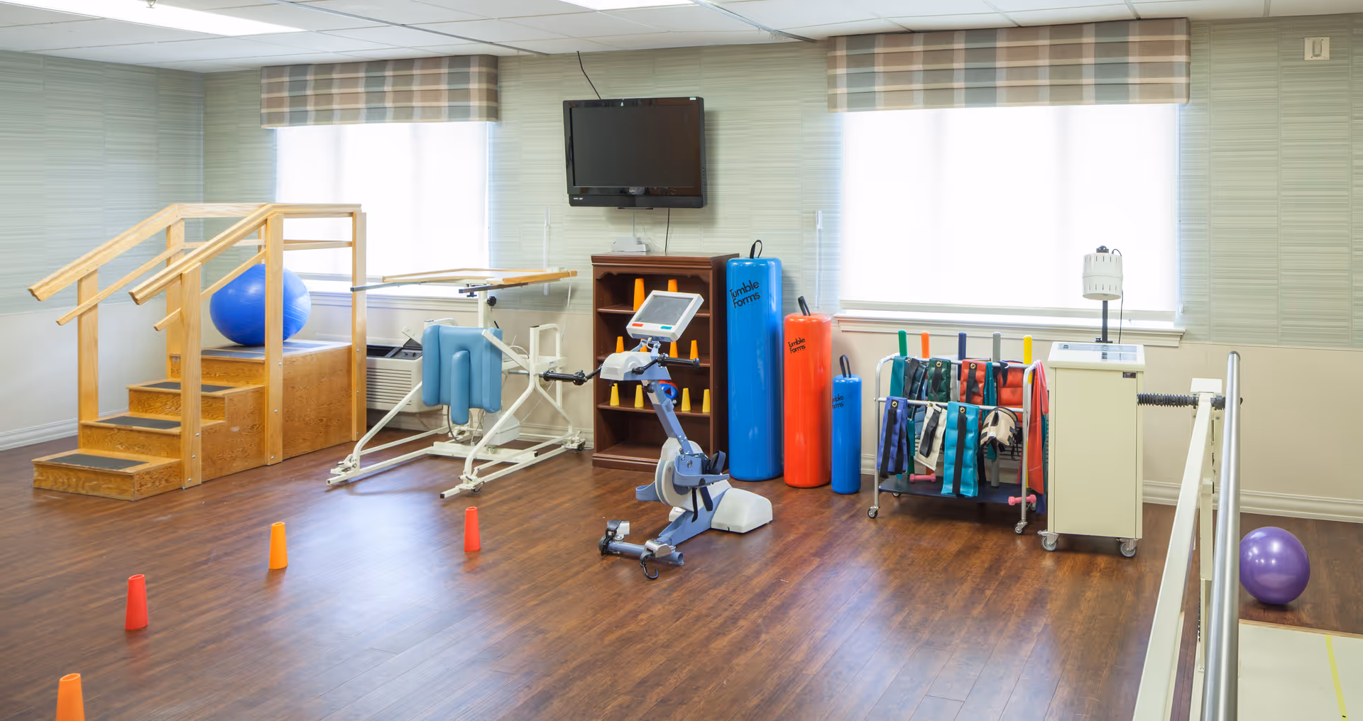 A physical therapy room with wooden stairs, exercise equipment including a stationary bike, therapy balls, resistance bands, and cones on a wooden floor. The room has two windows with plaid valances and a wall-mounted TV above a small wooden shelf.