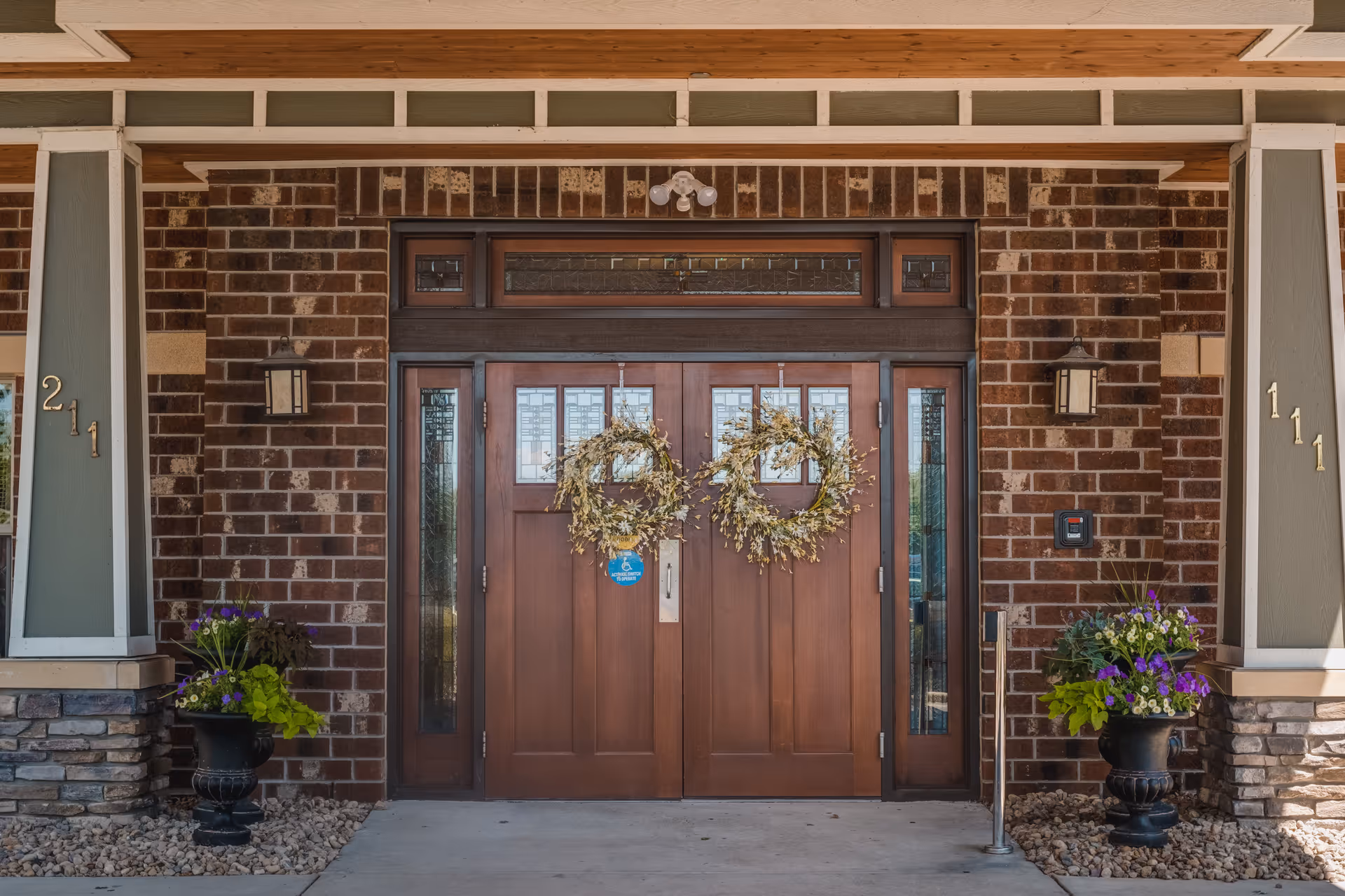 Front entrance of a building with double wooden doors decorated with two wreaths. The entrance is framed by brick walls and stone pillars with house numbers 211 and 111. Two black planters with green and purple flowers are placed on either side of the doors.