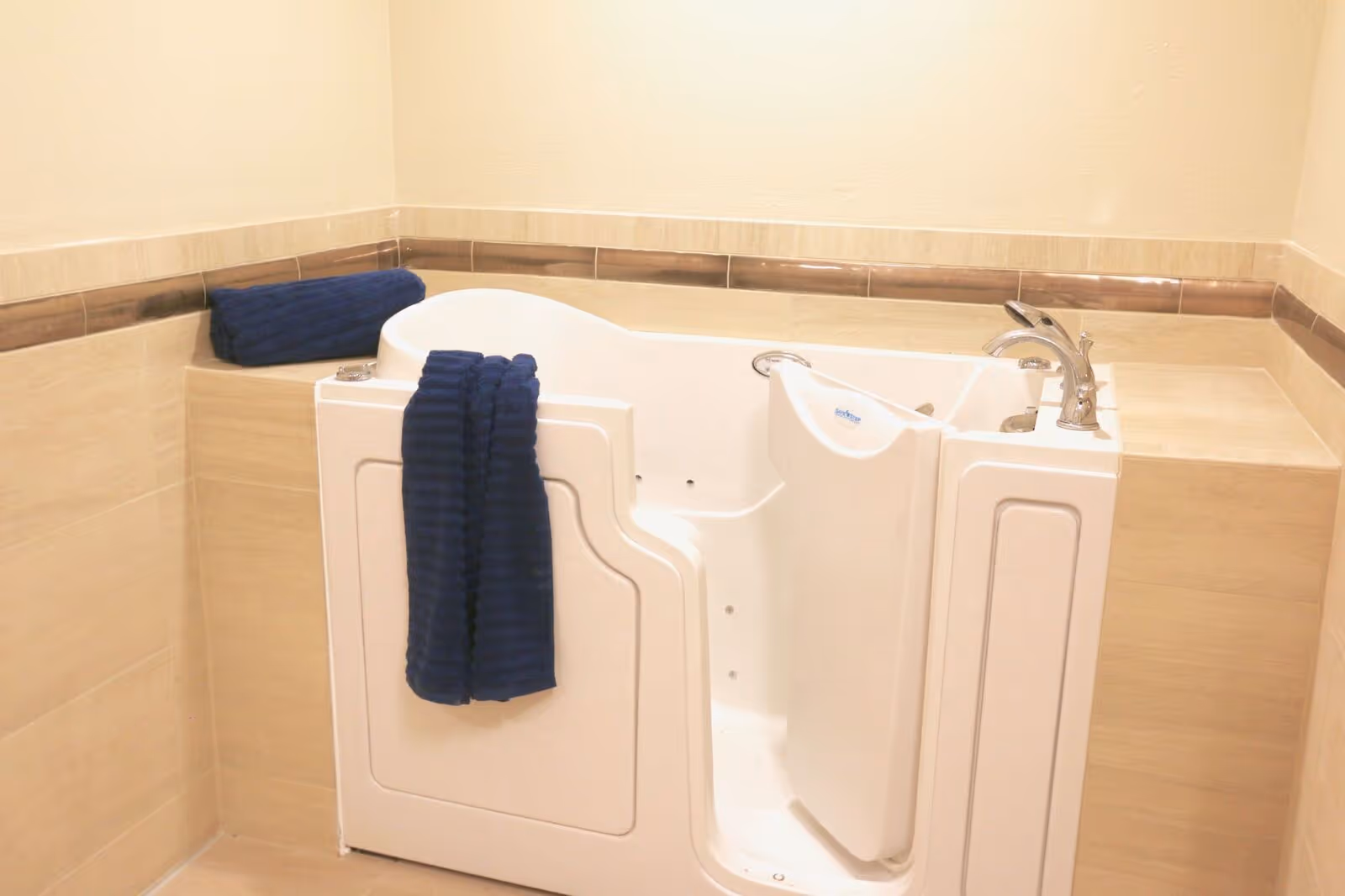 A walk-in bathtub with a door, faucet, and a blue towel hanging on the side, set against beige tiled walls with a decorative brown tile border.