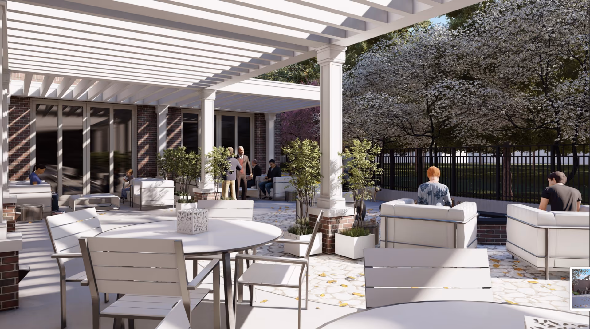 Outdoor patio area with white pergola overhead, white tables and chairs, potted plants, and several people sitting and standing. Trees with white blossoms and a black metal fence are visible in the background.
