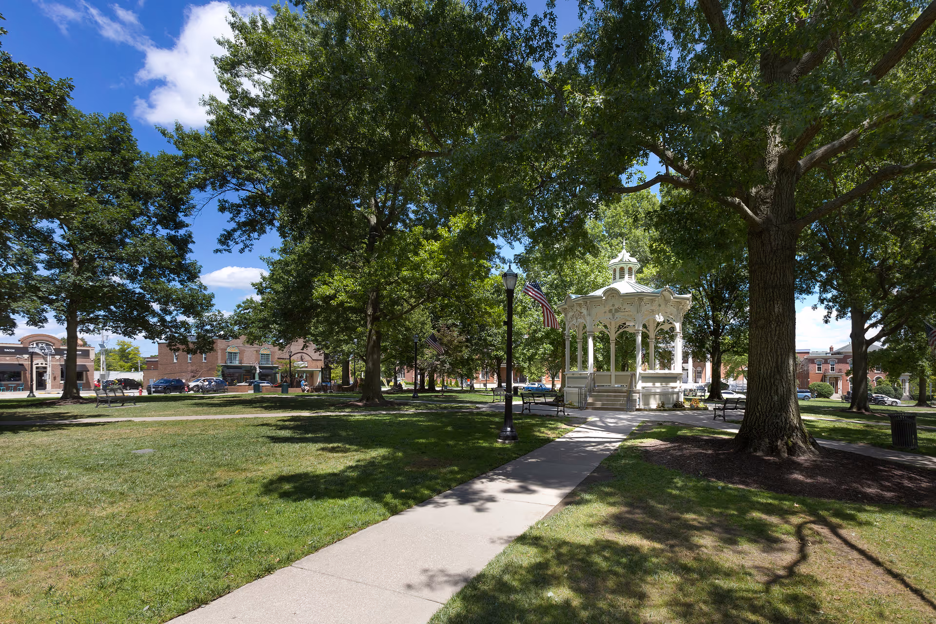 A sunny park scene with a paved walkway leading to a white gazebo surrounded by large green trees and grass. American flags are displayed on lamp posts along the path, and buildings are visible in the background under a blue sky with some clouds.