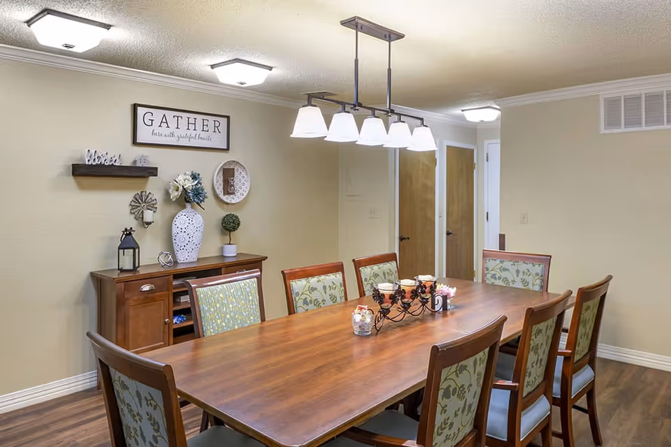 A dining room with a long wooden table surrounded by eight chairs with floral upholstery. Above the table hangs a modern light fixture with six lamps. Against the wall is a wooden sideboard decorated with a large white vase with flowers, a small topiary, a lantern, and other decorative items. On the wall above the sideboard is a sign that reads 'GATHER here with grateful hearts'. The room has beige walls, wooden doors, and wood flooring.