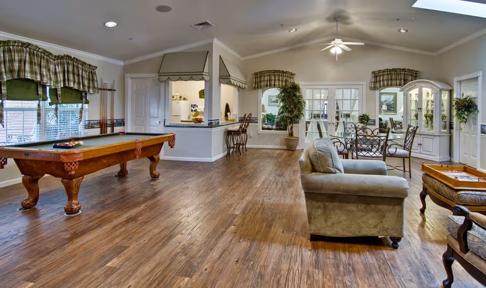 A spacious senior living common area featuring a pool table on the left, a cozy seating area with a sofa and chairs on the right, and a dining table with chairs near the back. The room has wooden flooring, large windows with green plaid curtains, potted plants, and a ceiling fan with lights.