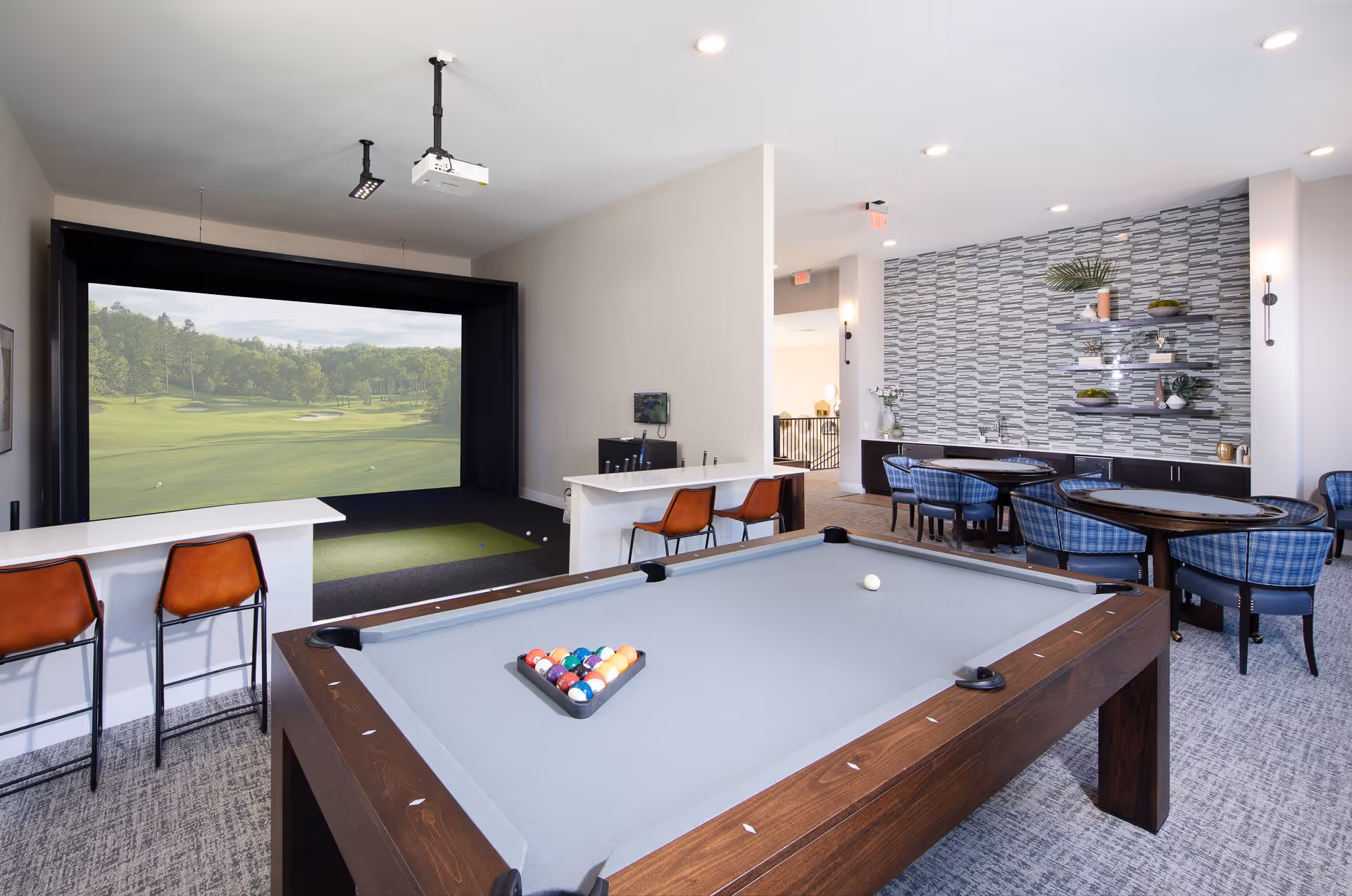 A recreational room featuring a pool table with a rack of billiard balls and a white cue ball, a golf simulator with a large screen displaying a golf course, two brown bar stools at a white counter, and a seating area with round tables and blue plaid chairs against a decorative tiled wall with shelves holding plants and decor items.