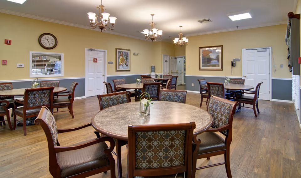 A dining room with multiple round tables and cushioned chairs arranged neatly. The room has wooden flooring, yellow and gray walls with white trim, framed artwork, a clock, and several chandeliers hanging from the ceiling. There is a water dispenser and a phone mounted on the wall near white doors.