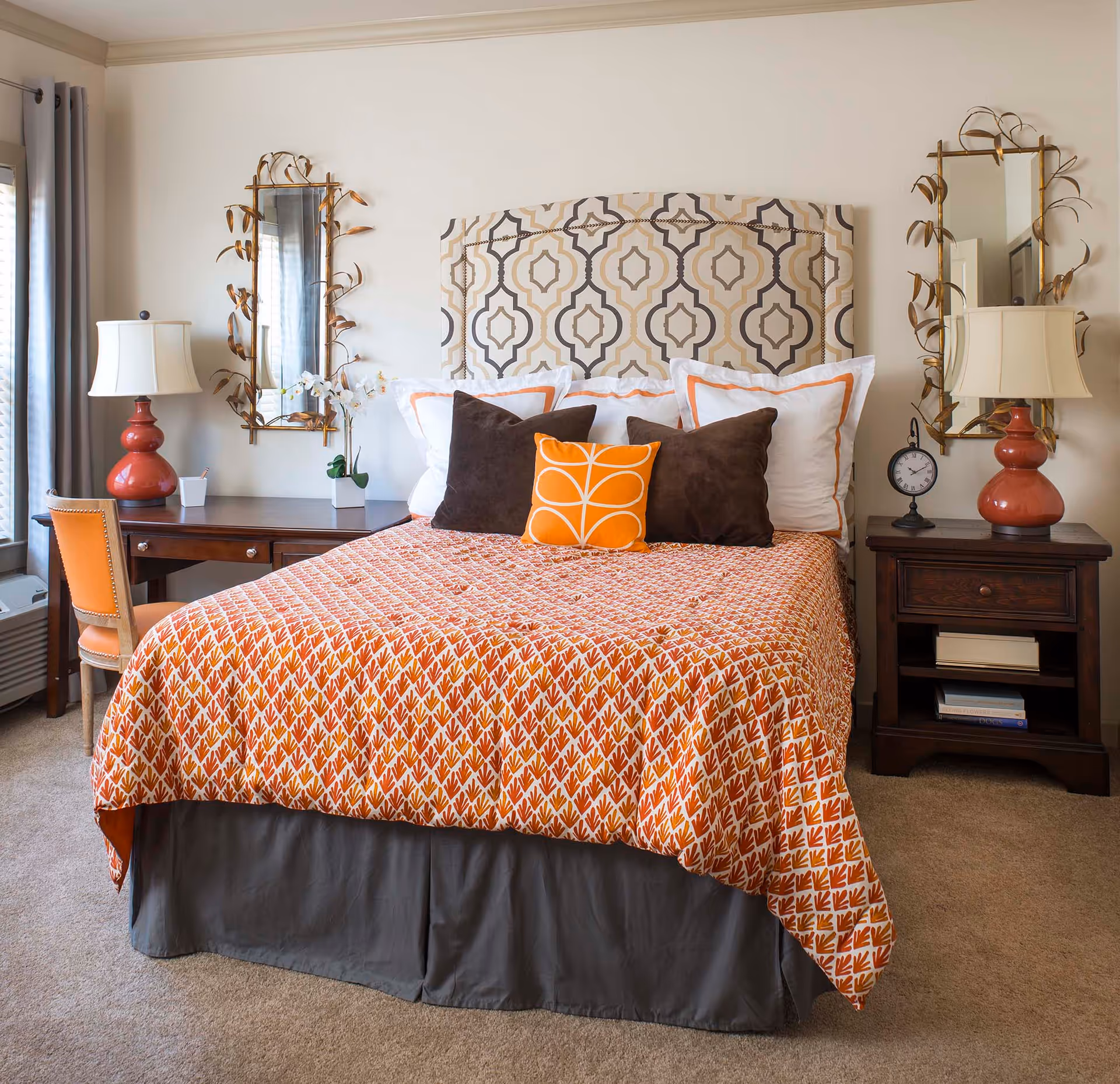 A neatly made bedroom with a patterned headboard, orange patterned bedspread and decorative pillows, flanked by wooden nightstands and lamps.