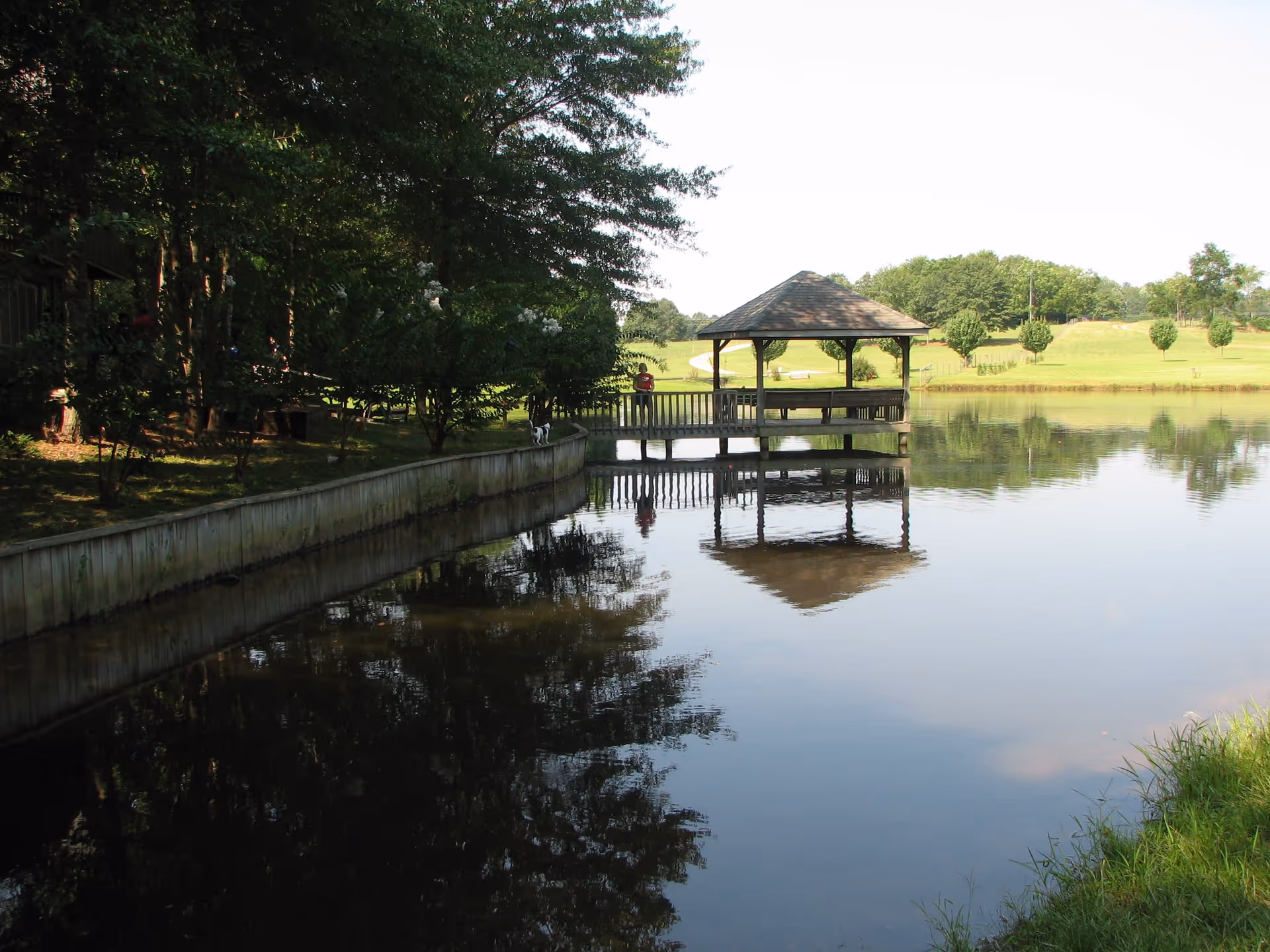 A peaceful outdoor scene at Lakeview Manor featuring a wooden gazebo on a pier extending over a calm pond. The pond reflects the gazebo and surrounding trees. There is a grassy area and trees in the background under a clear sky.