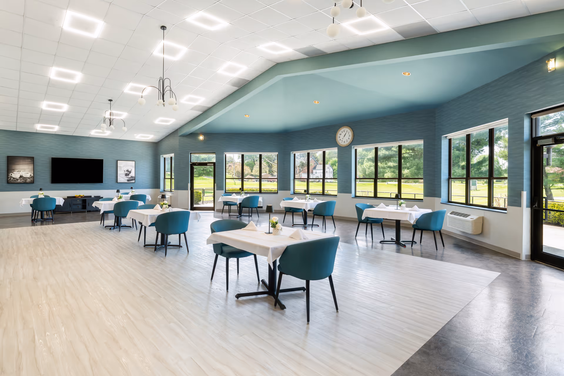 A spacious, well-lit dining room with several tables covered with white tablecloths and blue chairs. Large windows let in natural light and offer views of green outdoor scenery. The room has modern ceiling lights, a wall clock, and framed artwork on the walls.