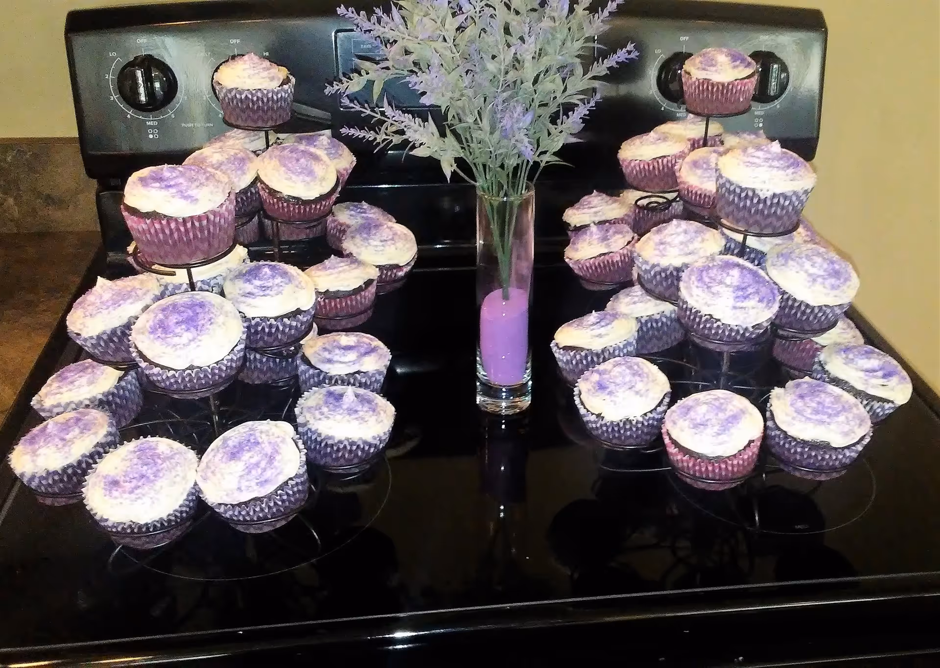 Two multi-tiered cupcake stands filled with cupcakes topped with white frosting and purple sprinkles, placed on a black stovetop. A tall clear vase with purple flowers and purple sand is positioned between the cupcake stands.