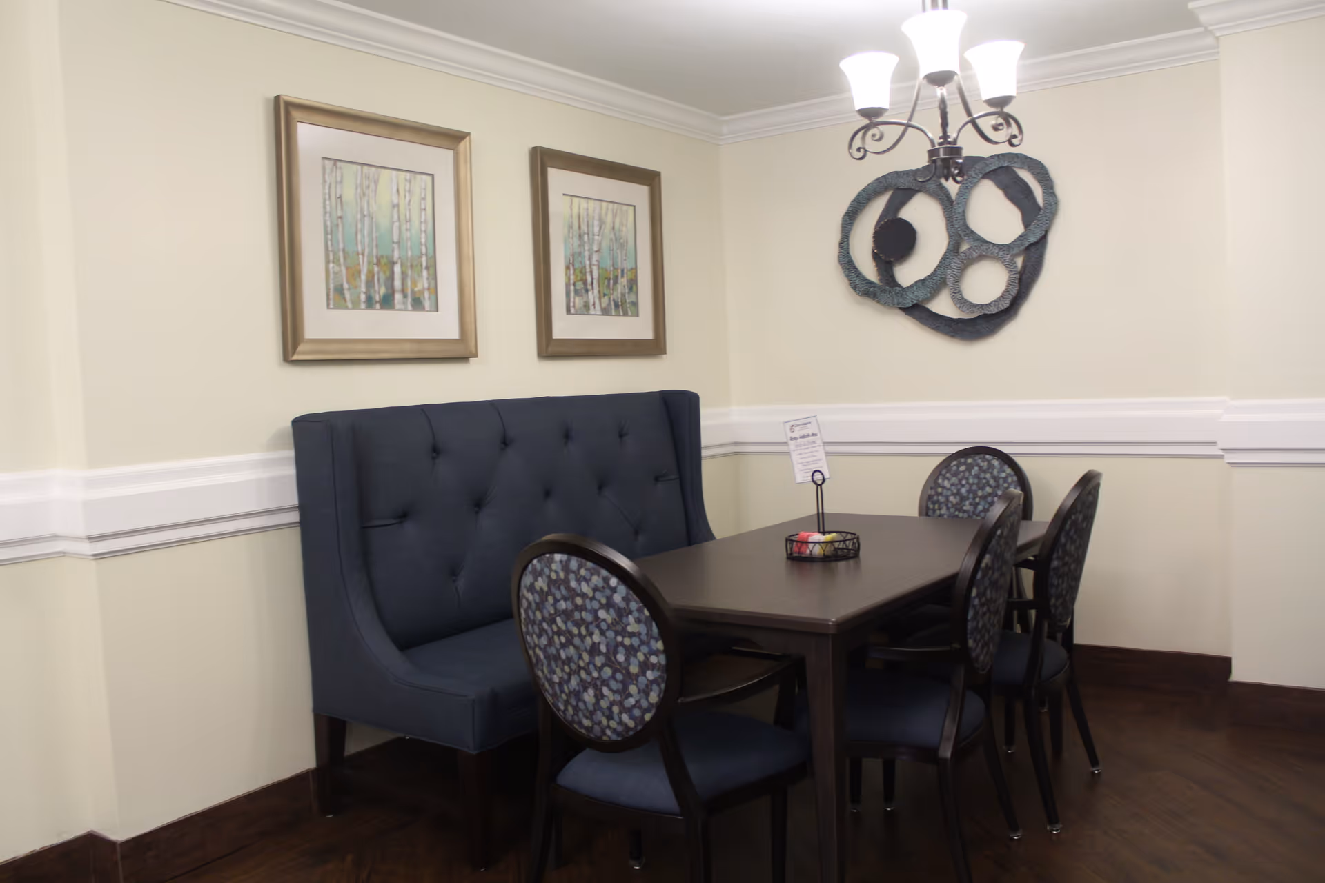 A dining area with a dark wooden table surrounded by four chairs and a blue upholstered bench. The chairs have patterned blue cushions. On the wall above the bench are two framed pictures of birch trees, and a decorative circular wall art piece is mounted on the adjacent wall. A chandelier with five lights hangs above the table.