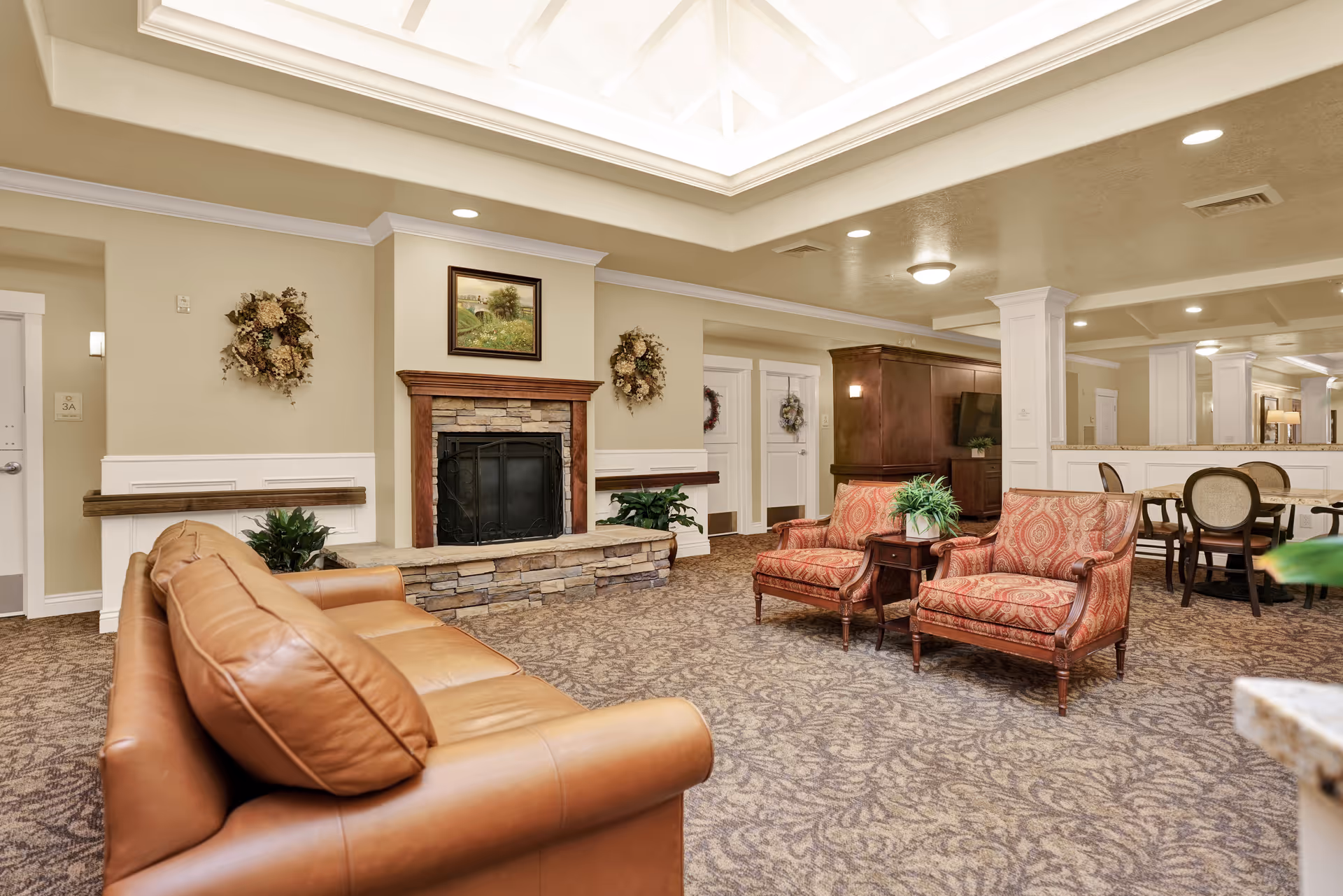 A cozy living room area in a senior living facility featuring a brown leather sofa, two patterned armchairs, a stone fireplace with a wooden mantle, decorative wreaths on the walls, and a dining area with chairs and tables in the background under soft lighting.