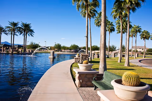 A curved walkway beside a large pond with water fountains, lined with palm trees and green benches. Large potted cacti are placed along the walkway, and there is a clear blue sky above.