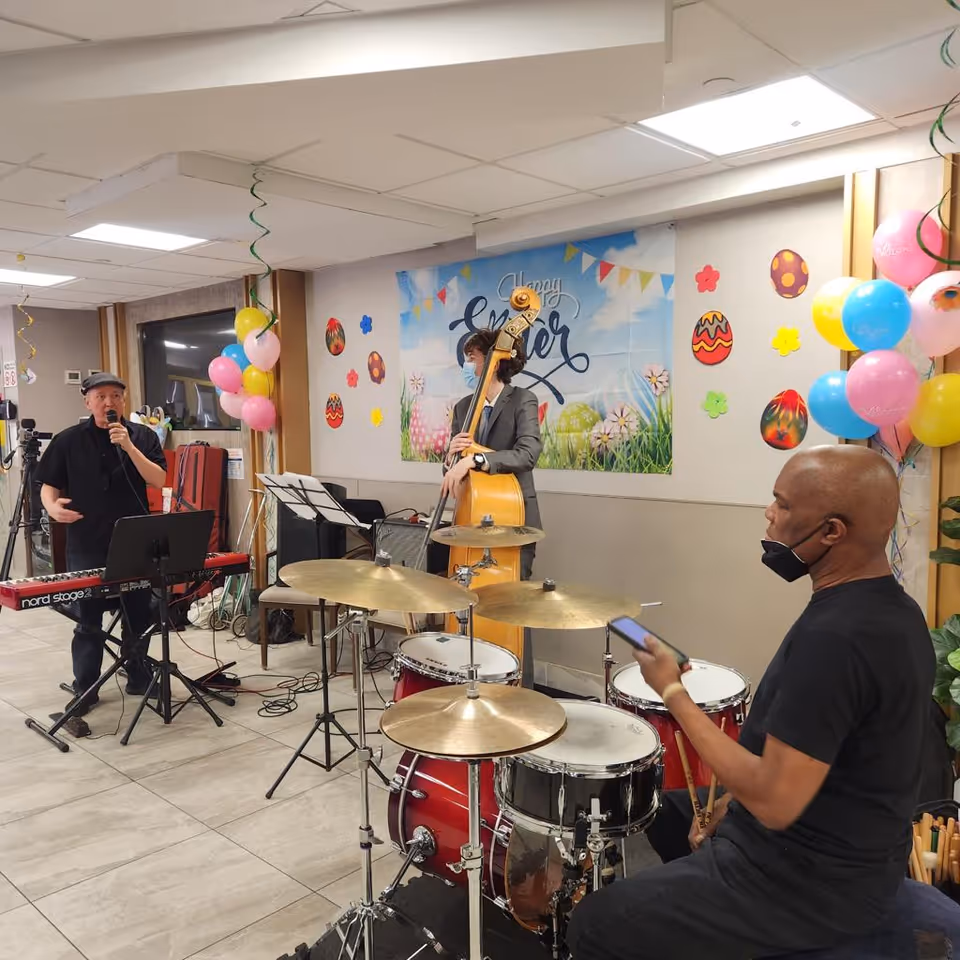 A small musical band performing indoors at a nursing home decorated for Easter. The band includes a keyboard player singing into a microphone, a double bass player wearing a mask, and a drummer holding drumsticks and looking at a phone. The room is decorated with colorful balloons and Easter-themed wall decorations including painted eggs and a banner that says 'Happy Easter'.