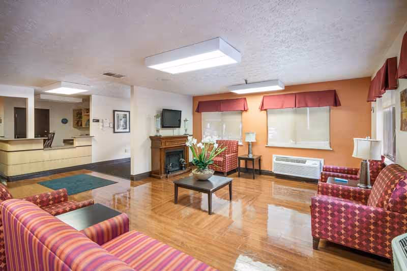 A spacious living room area with polished wooden floors, featuring patterned armchairs and a sofa in shades of red and purple. There is a small coffee table with a flower arrangement in the center, a fireplace with a TV mounted above it, and windows with red valances. The room is well-lit with ceiling lights and has a reception desk visible in the background.
