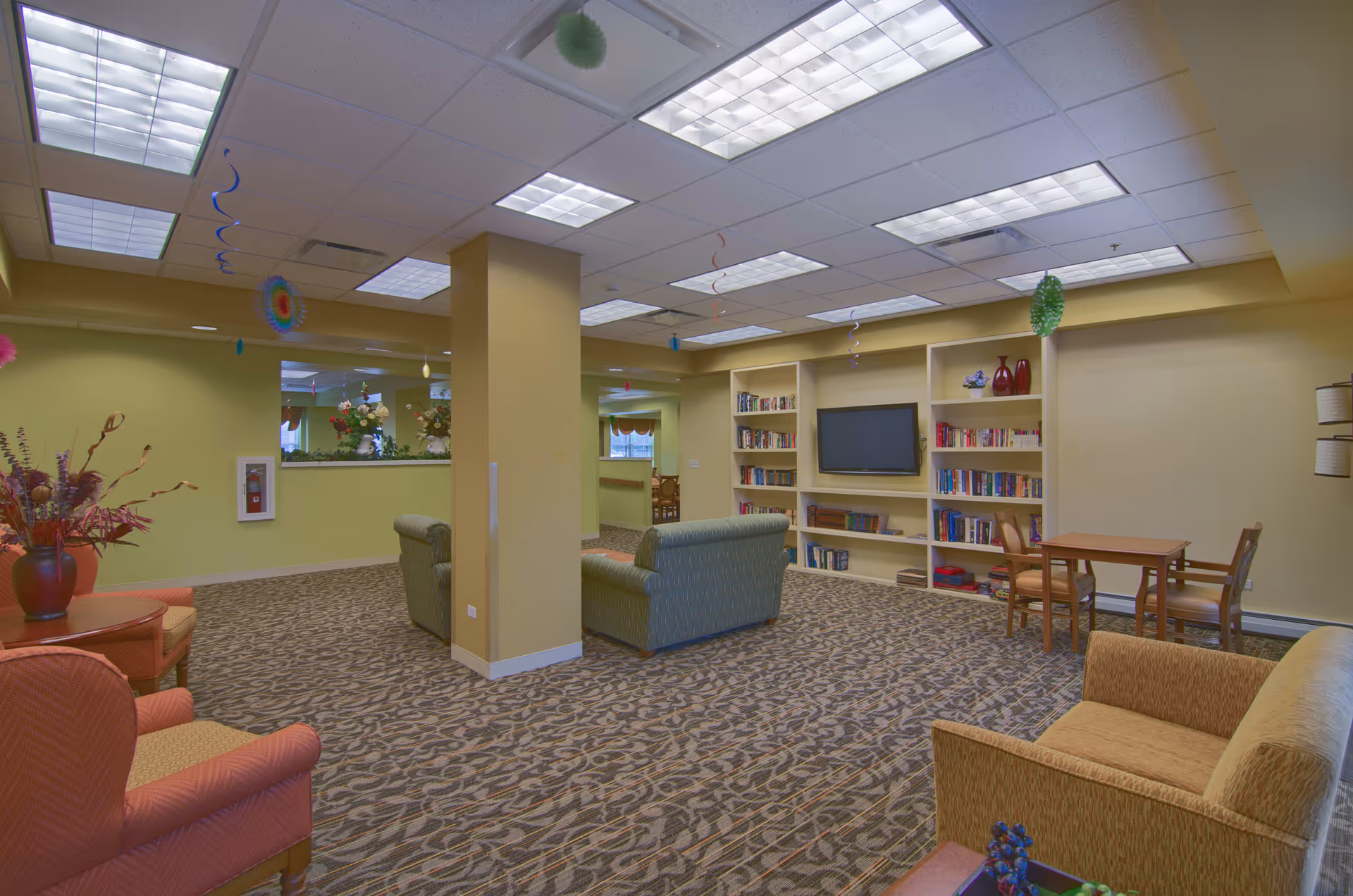 A spacious senior living facility common area with patterned carpet, multiple armchairs and sofas, a small table with chairs, built-in bookshelves filled with books, and a wall-mounted flat-screen TV. The ceiling has recessed lighting and colorful hanging decorations.