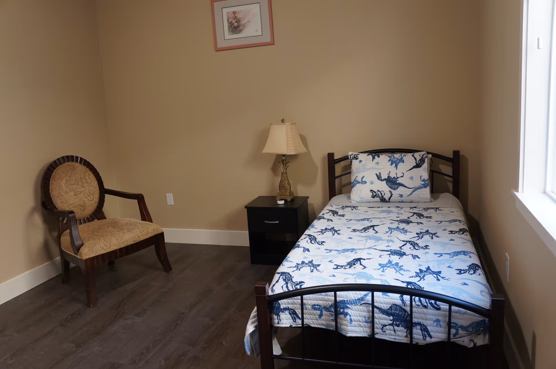 Small bedroom with a single metal-framed bed covered in blue patterned bedding, a nightstand with a lamp, and an upholstered chair.