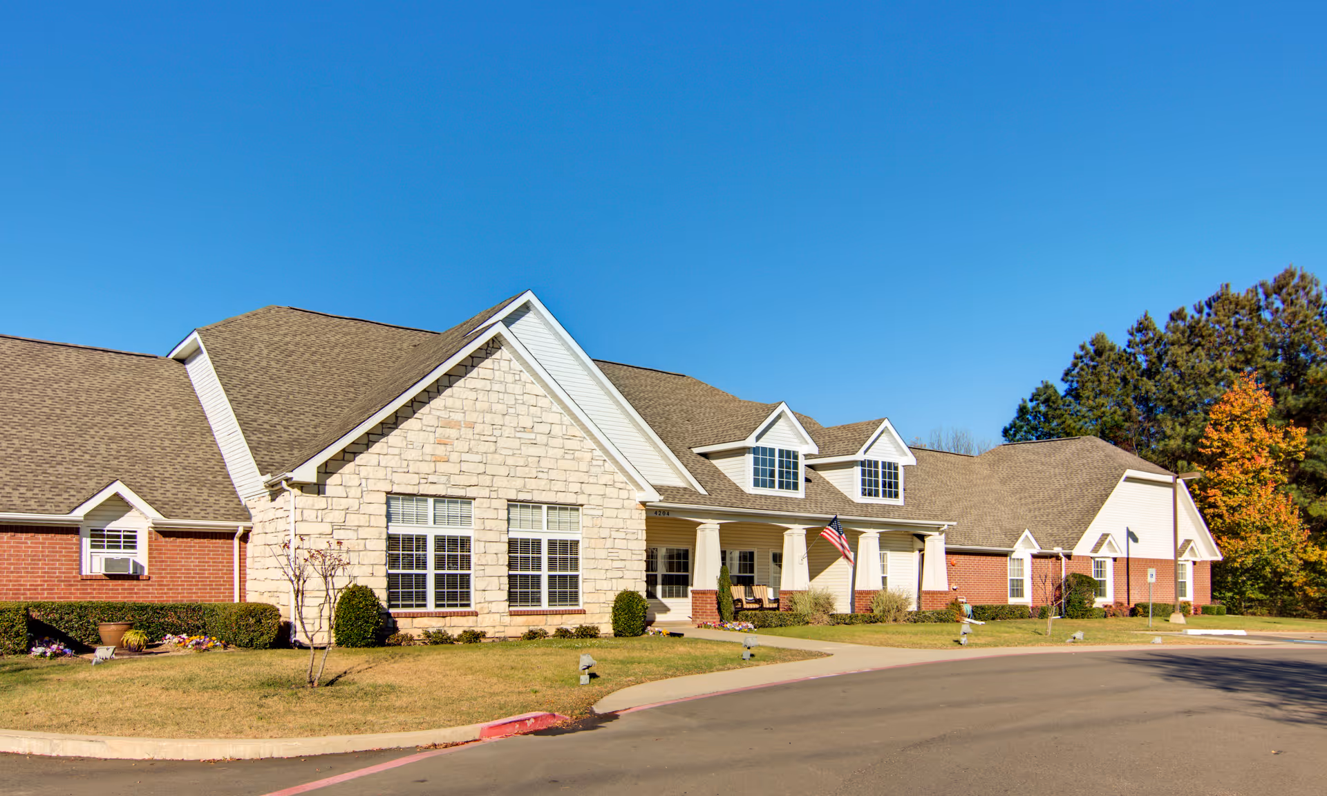 Exterior view of a single-story senior living facility building with a combination of brick and stone facade, multiple windows, a covered porch with chairs, an American flag, and a clear blue sky above. The surrounding area includes a paved driveway, manicured lawn, shrubs, and trees with some autumn foliage.