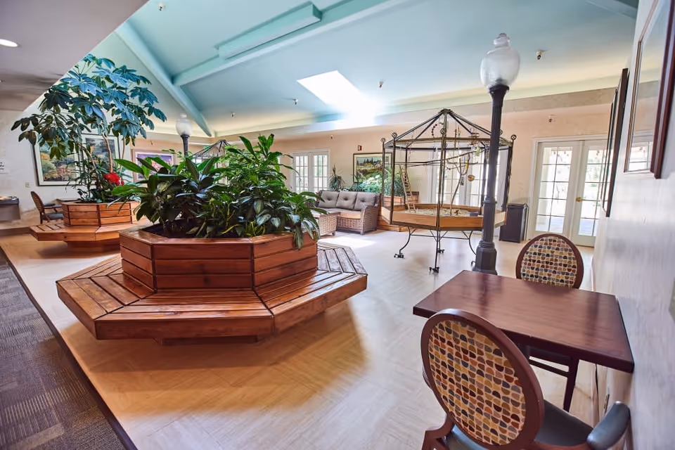 A bright and spacious indoor common area with large wooden planters filled with green plants, a skylight in the ceiling, cushioned seating, a birdcage, and a small table with two patterned chairs. The room has light-colored flooring and walls, with framed artwork and glass doors leading outside.