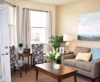 A cozy living room with a beige sofa adorned with yellow and blue pillows, a wooden coffee table with a flower arrangement, two patterned chairs near a window with light curtains, a side table with a lamp and framed photo, and a large abstract painting on the wall.