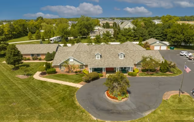 Aerial view of a single-story senior living facility named Caregivers Inn surrounded by well-maintained lawns, trees, and shrubs. The building has a gray shingled roof with dormer windows and a circular driveway with a landscaped island in the center. Several cars are parked in the parking area to the right, and an American flag is visible near the driveway.