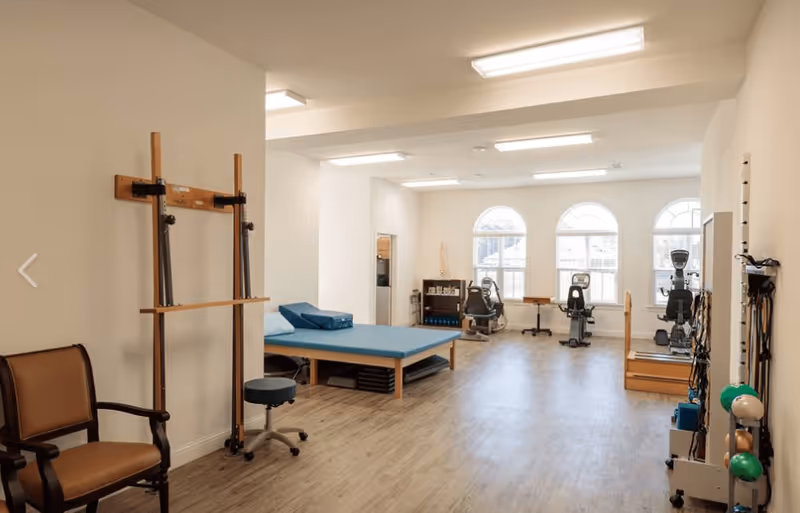 A spacious physical therapy or rehabilitation room with exercise equipment including stationary bikes, a padded therapy table, resistance bands, and various exercise balls. The room has large arched windows letting in natural light and wood-style flooring.