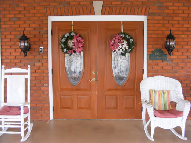 Front entrance of a building with double wooden doors decorated with floral wreaths. The doors have oval glass panels. On either side of the doors are two white chairs with cushions, one rocking chair and one wicker chair with a striped pillow. The exterior wall is made of red brick and has two black lantern-style wall lights. A green address plaque with the number 1513 and the text 'Crest Pine Run' is mounted on the right side of the door.