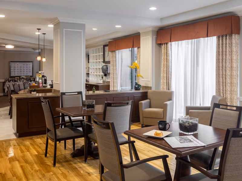 A well-lit common area in a senior living facility featuring wooden tables and cushioned chairs. There are two armchairs near large windows with sheer curtains and valances. On one table, there is a plate with a pastry, a coffee mug, and a newspaper. The room has polished wooden floors and a counter area with cabinets and decorative items, including a vase with yellow flowers.