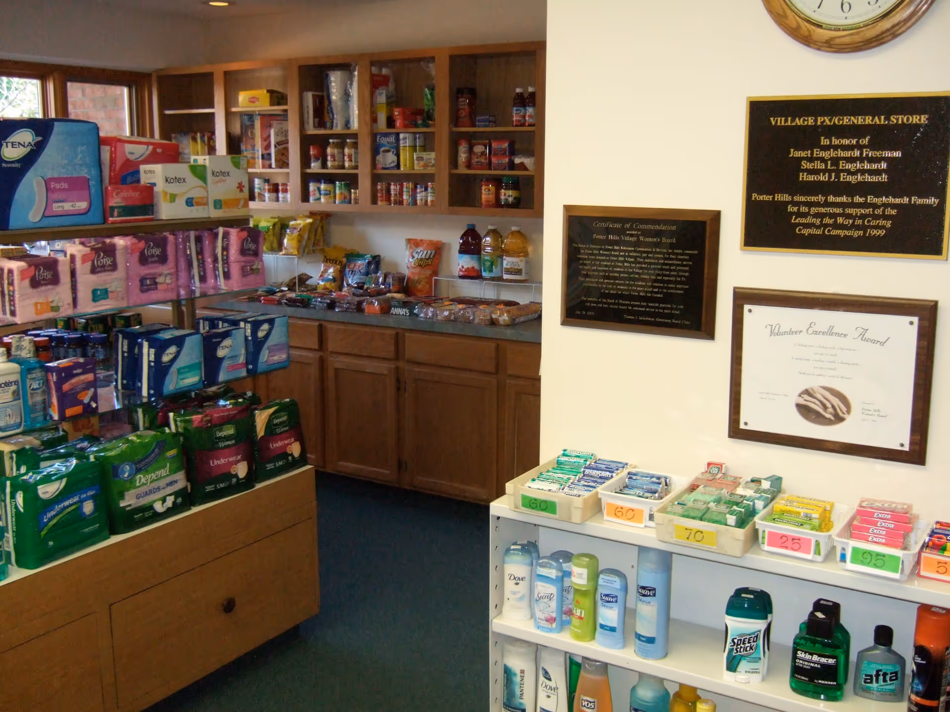 Interior view of a small general store or convenience area within a facility, displaying shelves stocked with personal care products, snacks, and hygiene items. There are wooden cabinets and shelves filled with various packaged goods, and plaques and awards are mounted on the wall.