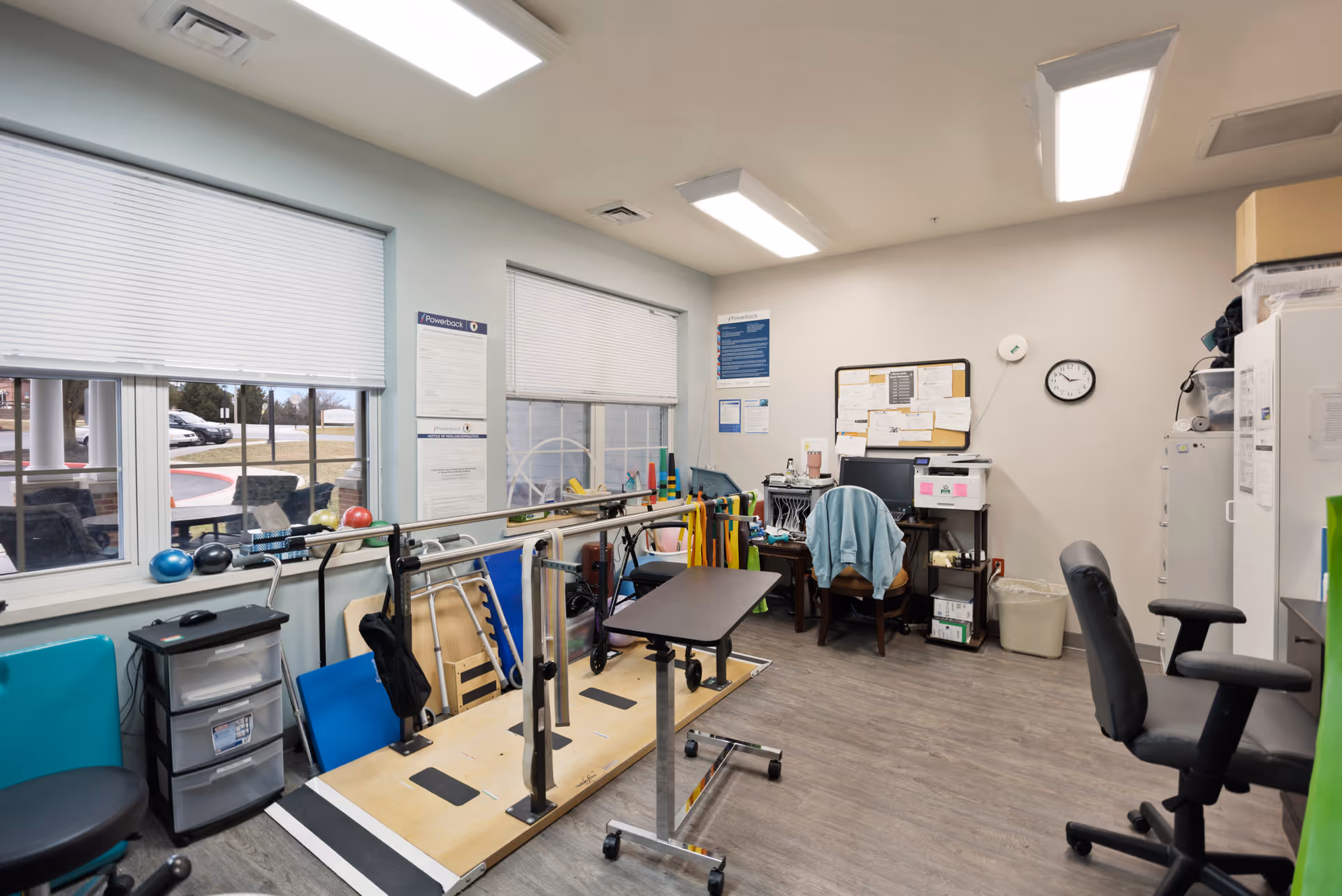 Rehabilitation therapy room with parallel walking bars, exercise equipment, a desk and chairs.
