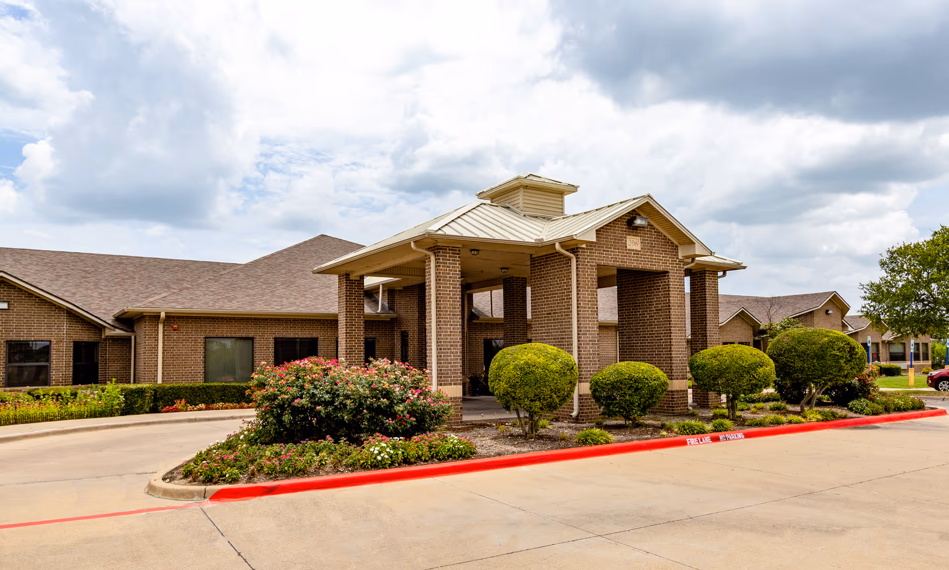 Exterior view of a single-story brick building with a covered entrance supported by brick columns. The building is surrounded by neatly trimmed bushes and flowering plants, with a driveway and red-painted curb in front. The sky is partly cloudy.