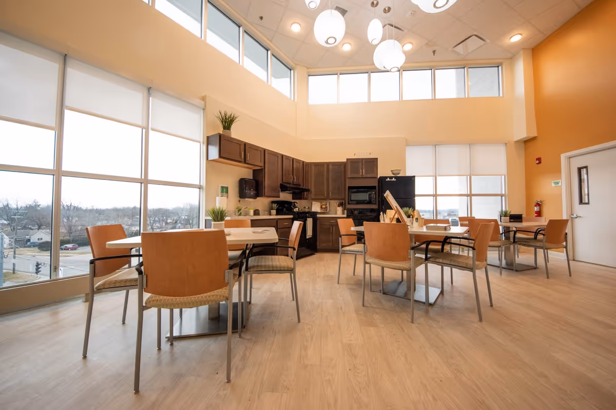 Bright and spacious senior living dining area with large windows, wooden tables and chairs, and a kitchen in the background featuring dark wood cabinets, a refrigerator, microwave, and stove. The room has light wood flooring and modern pendant lights hanging from the ceiling.