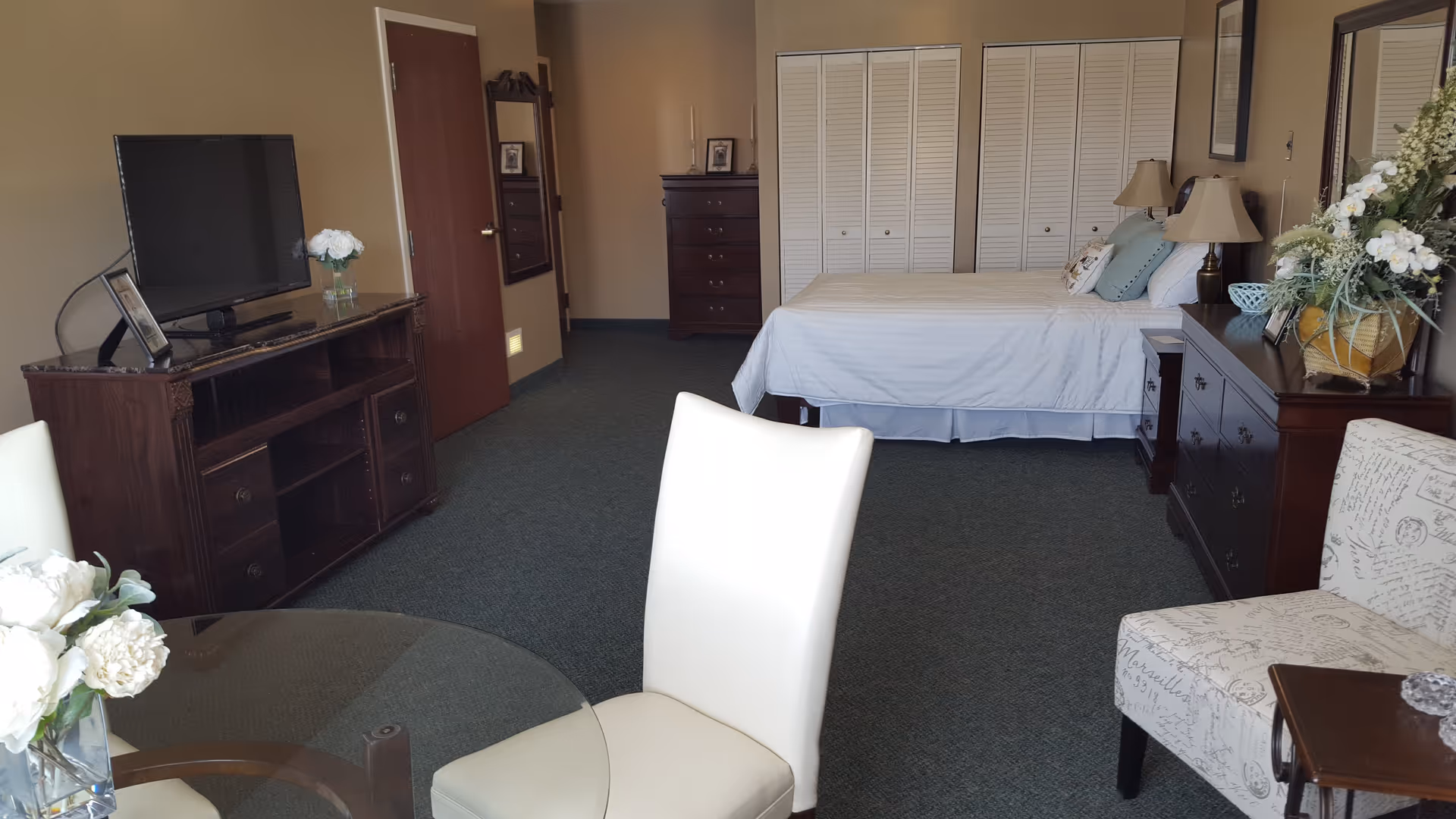A well-furnished bedroom in a senior living facility featuring a bed with white linens, two bedside tables with lamps, a dresser with a large floral arrangement, a chest of drawers, a TV on a wooden stand, a round glass table with white chairs, and a cushioned armchair with script pattern upholstery.