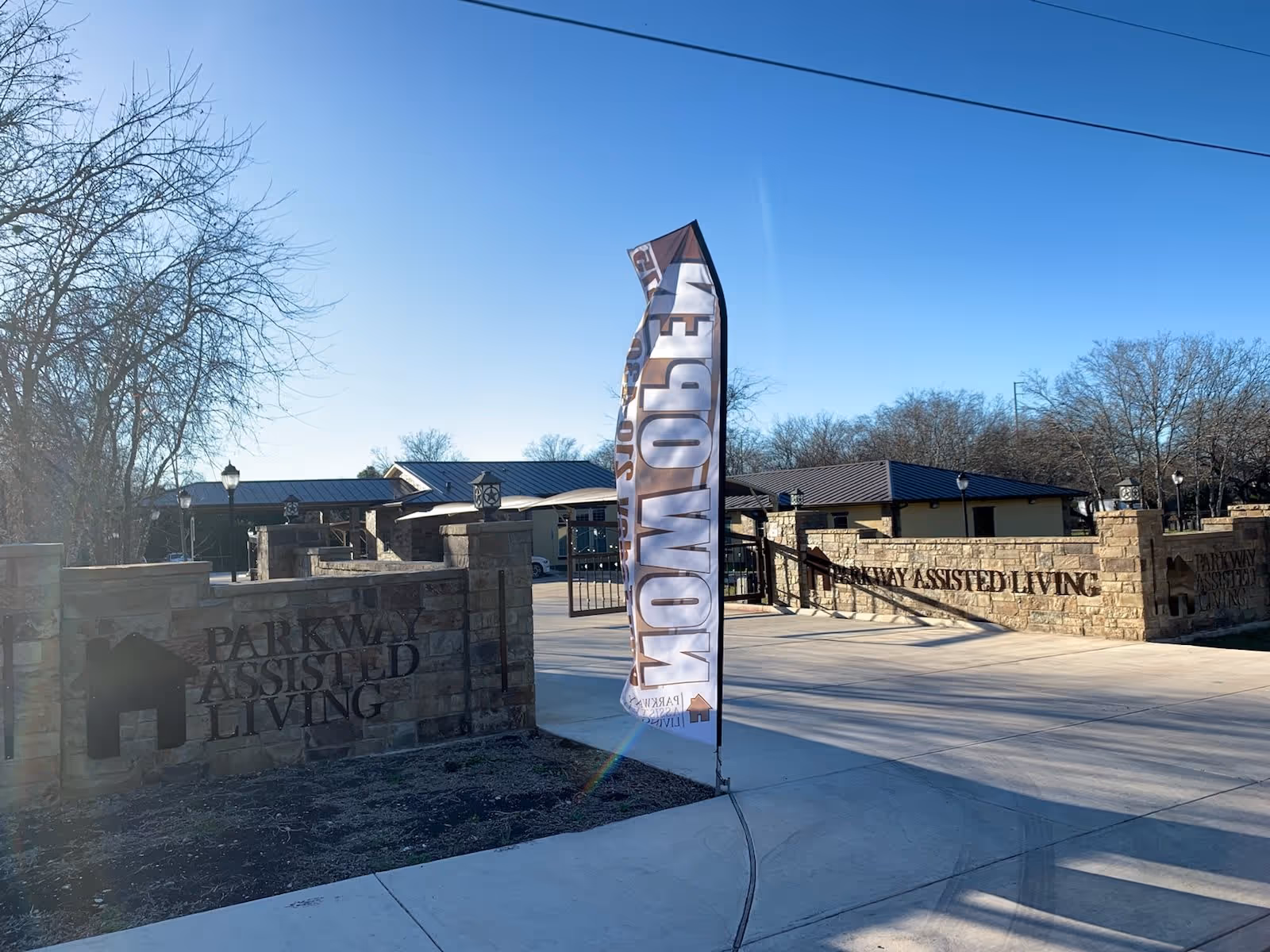 Entrance to Parkway Assisted Living facility with stone walls and signage on both sides of the driveway, a tall flag with the word 'NOW OPEN' fluttering in the breeze, and a clear blue sky above.