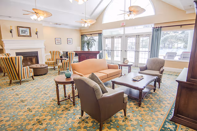 A bright and spacious living room area with a patterned carpet, a peach-colored sofa, two brown armchairs, a wooden coffee table, and side tables with decorative items. The room features large windows with curtains, a fireplace with framed pictures above it, and ceiling fans with light fixtures.