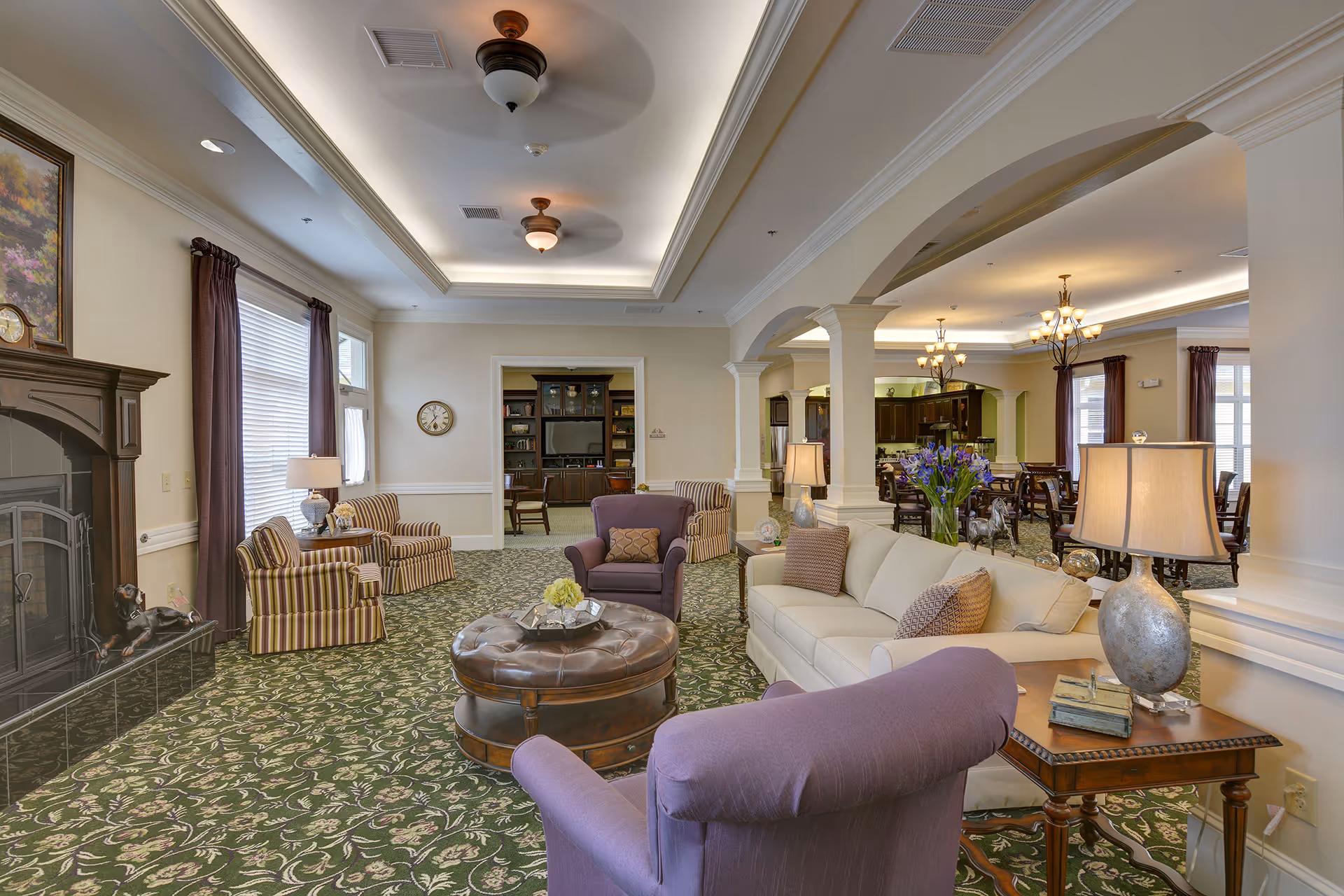 A spacious and well-lit living room area in a senior living facility featuring comfortable seating including a beige sofa, purple armchairs, and striped chairs. There is a round leather ottoman in the center, a fireplace with a dog statue on the hearth, and a side table with a lamp and decorative items. The room has patterned green carpet, large windows with curtains, ceiling fans, and an open archway leading to a dining area with tables and chairs.