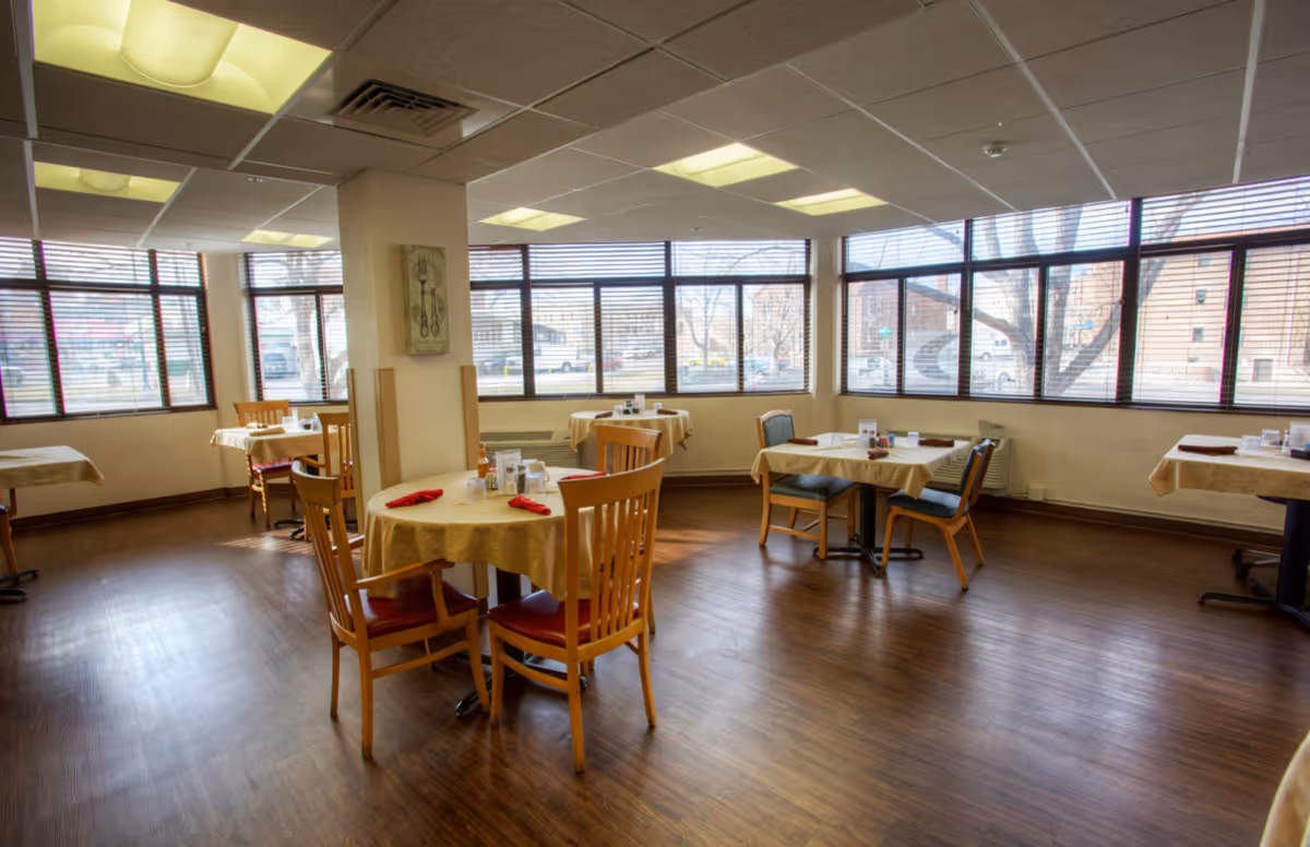 A dining room with several round tables covered with beige tablecloths, each set with red napkins and condiments. The room has large windows with blinds allowing natural light to enter, wooden chairs with red and blue cushions, and a wooden floor.