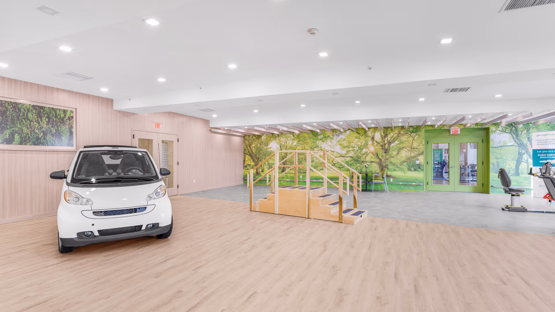 A spacious rehabilitation room with light wood flooring and a small white car positioned on the left side. In the center, there is a wooden staircase with handrails designed for physical therapy. The back wall features a large mural of a green park with trees and sunlight. To the right, there is exercise equipment and a green double door exit. The room is brightly lit with recessed ceiling lights.