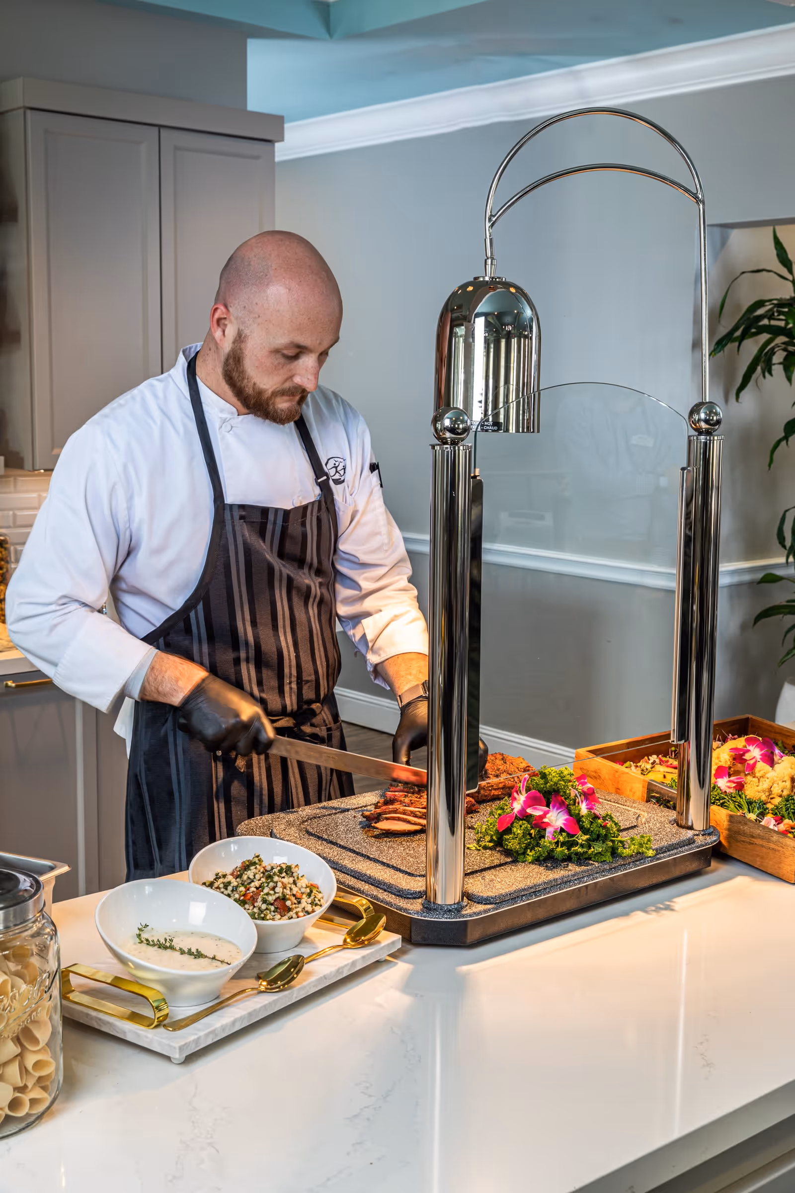 A chef wearing a white coat and black apron is slicing meat on a carving station with a heat lamp. There are bowls of side dishes and garnishes on the counter, and the setting appears to be a kitchen or food service area.