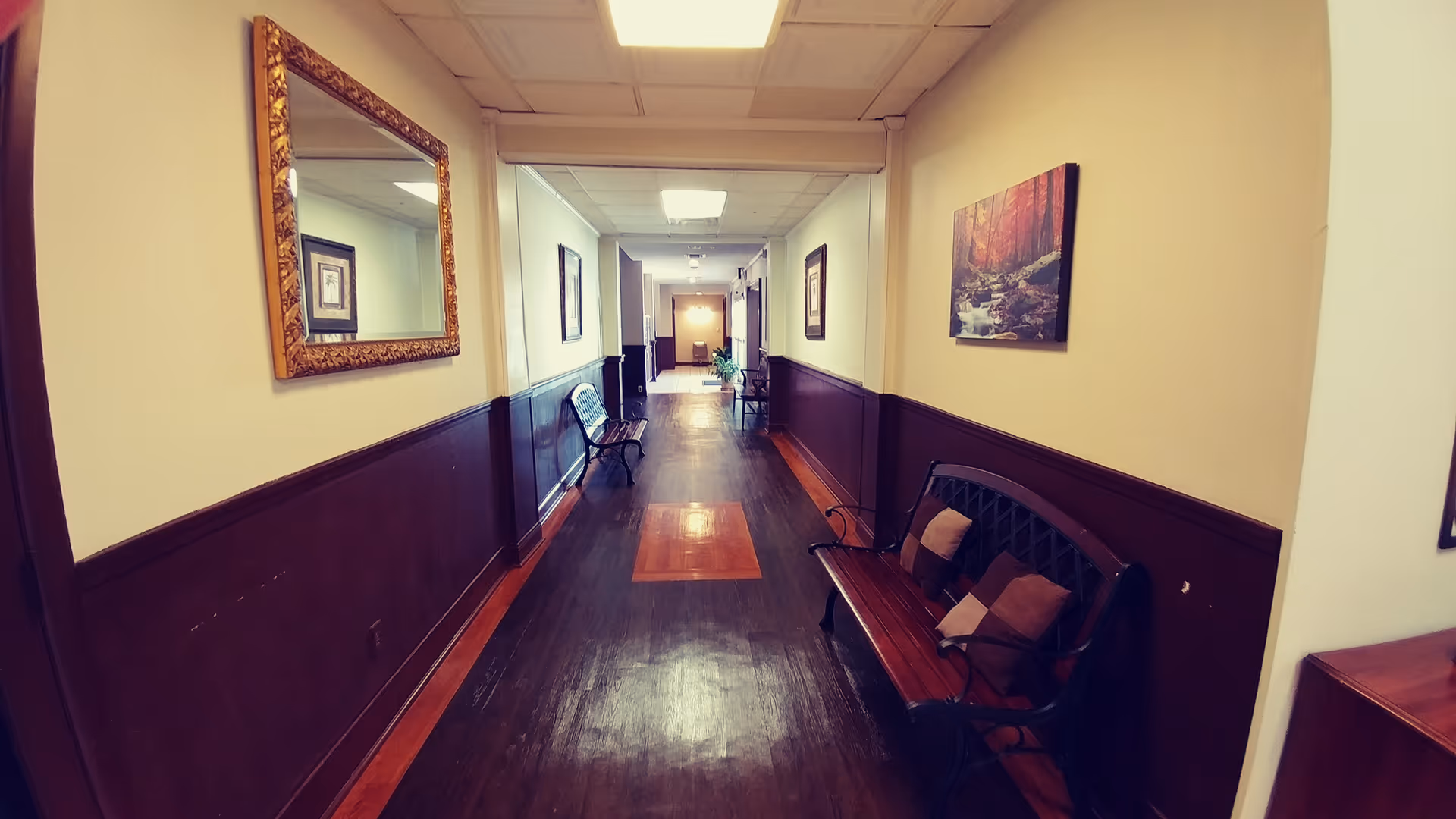 A long indoor hallway with dark wooden flooring and cream-colored walls with dark brown wainscoting. There are benches with cushions along the walls and framed artwork hanging above them. A large ornate mirror is mounted on the left wall. The hallway is well-lit with ceiling lights and leads to a door at the far end.