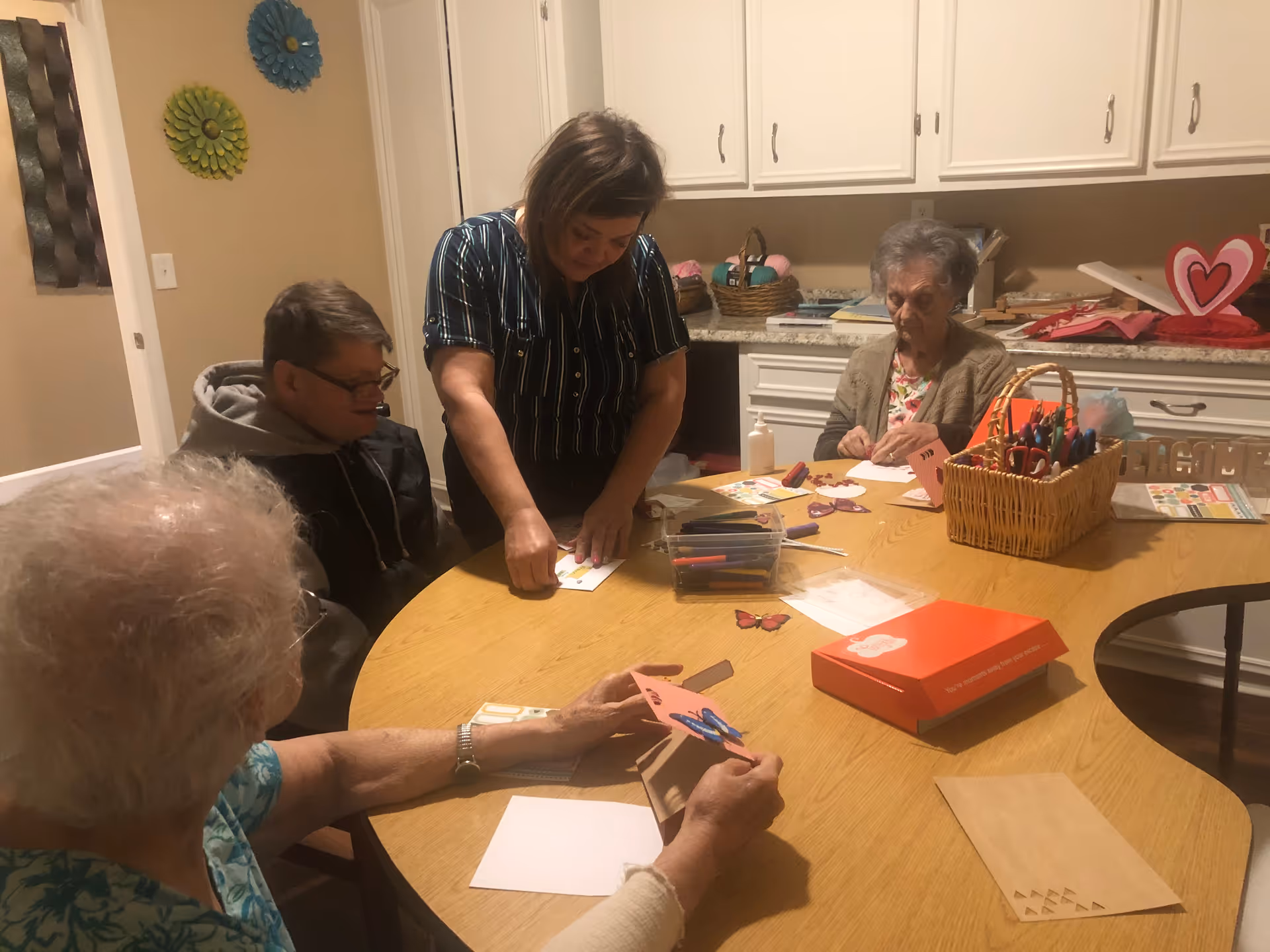 Four elderly individuals and a caregiver sitting around a wooden table engaged in a crafting activity with paper, markers, and scissors in a room with white cabinets and decorative wall hangings.