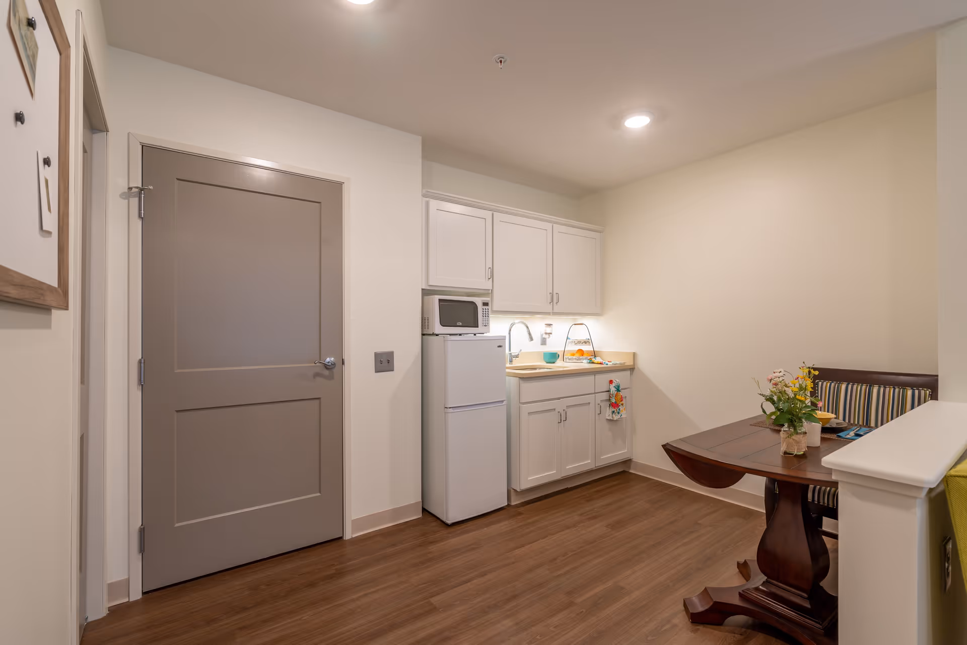 Small kitchen area with white cabinets, a microwave on top of a white refrigerator, a sink with a countertop, and a wooden dining table with a striped cushioned bench. The room has wood flooring and beige walls with a closed gray door on the left.